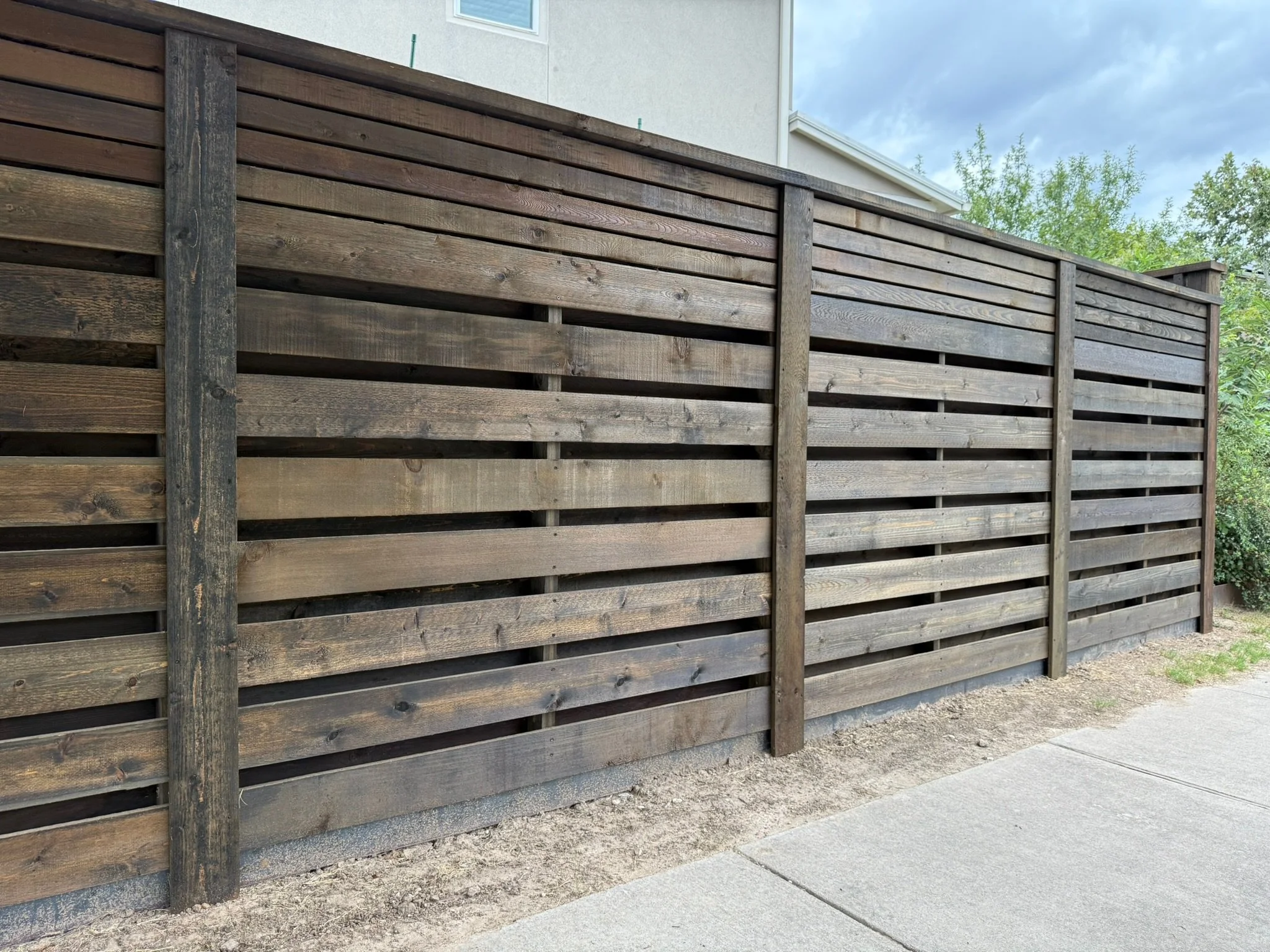 A horizontal wooden privacy fence with burnt hickory stain and vertical support posts, located along a concrete sidewalk next to a house with a white exterior. Trees and cloudy sky are visible in the background.