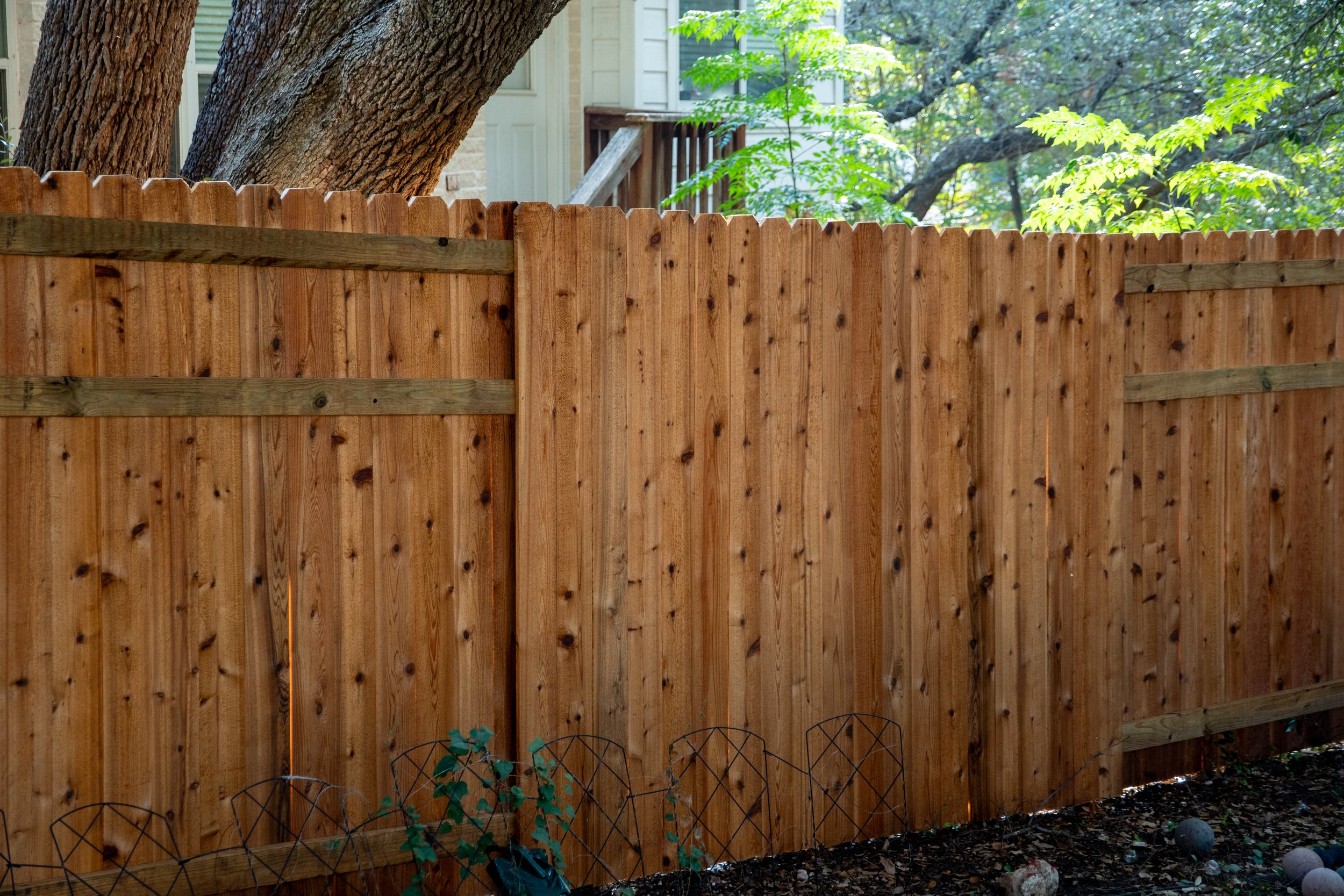Newly installed wooden privacy fence in backyard with a large tree and a house with a deck in the background.