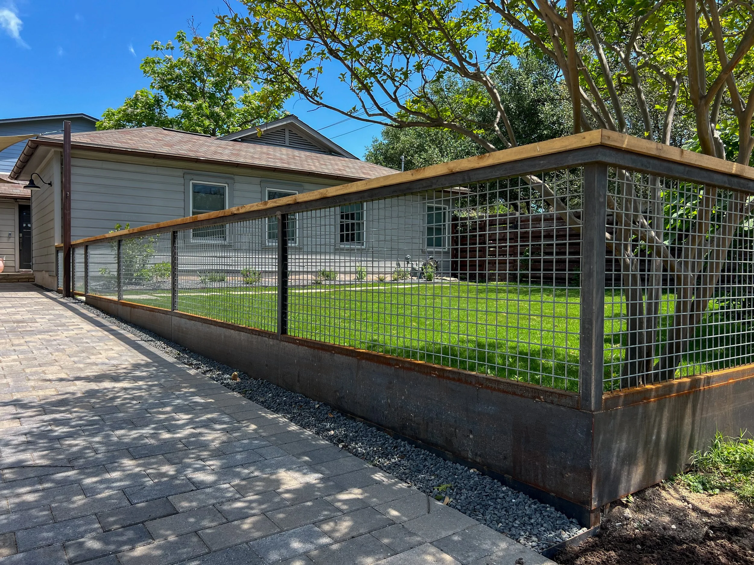 A backyard with a wooden and metal wire fence, a well-maintained green lawn, a tree with multiple branches, and a house with a gray exterior and windows, under a clear blue sky.