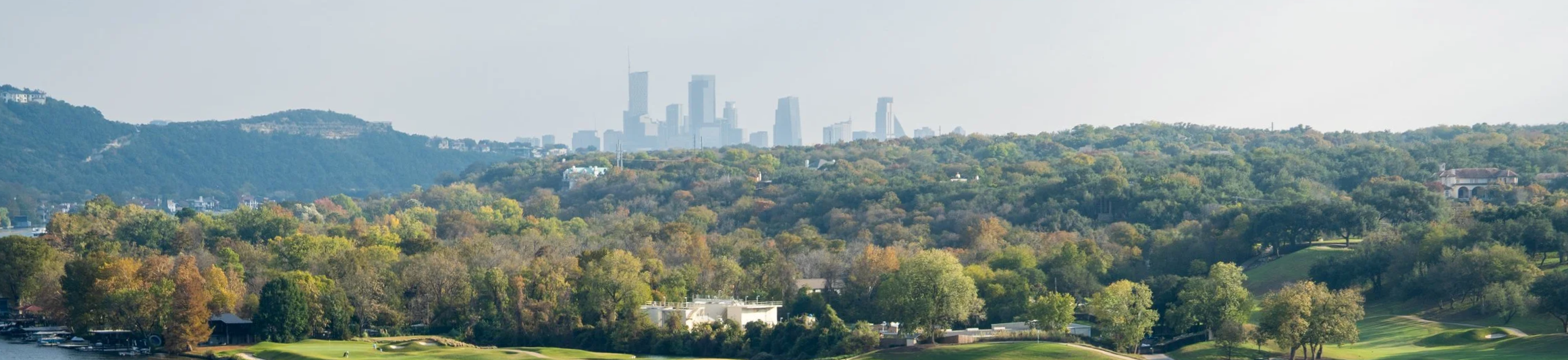 A scenic view of green trees in a hilly area with a city skyline featuring tall buildings in the background.