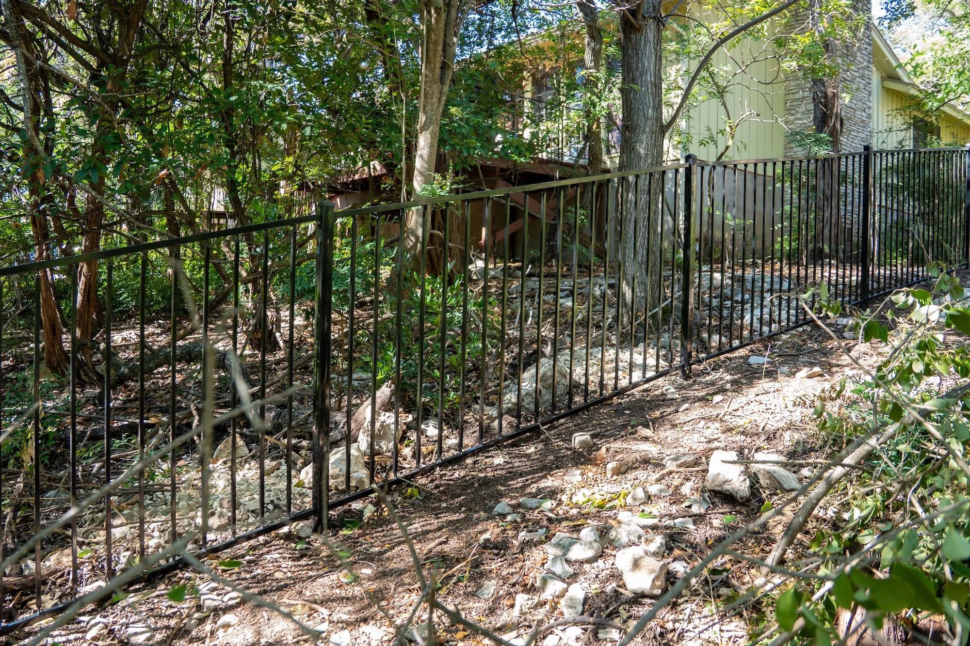 Black metal fence on a dirt and rocky ground with trees and bushes in a wooded backyard, with part of a house visible in the background.