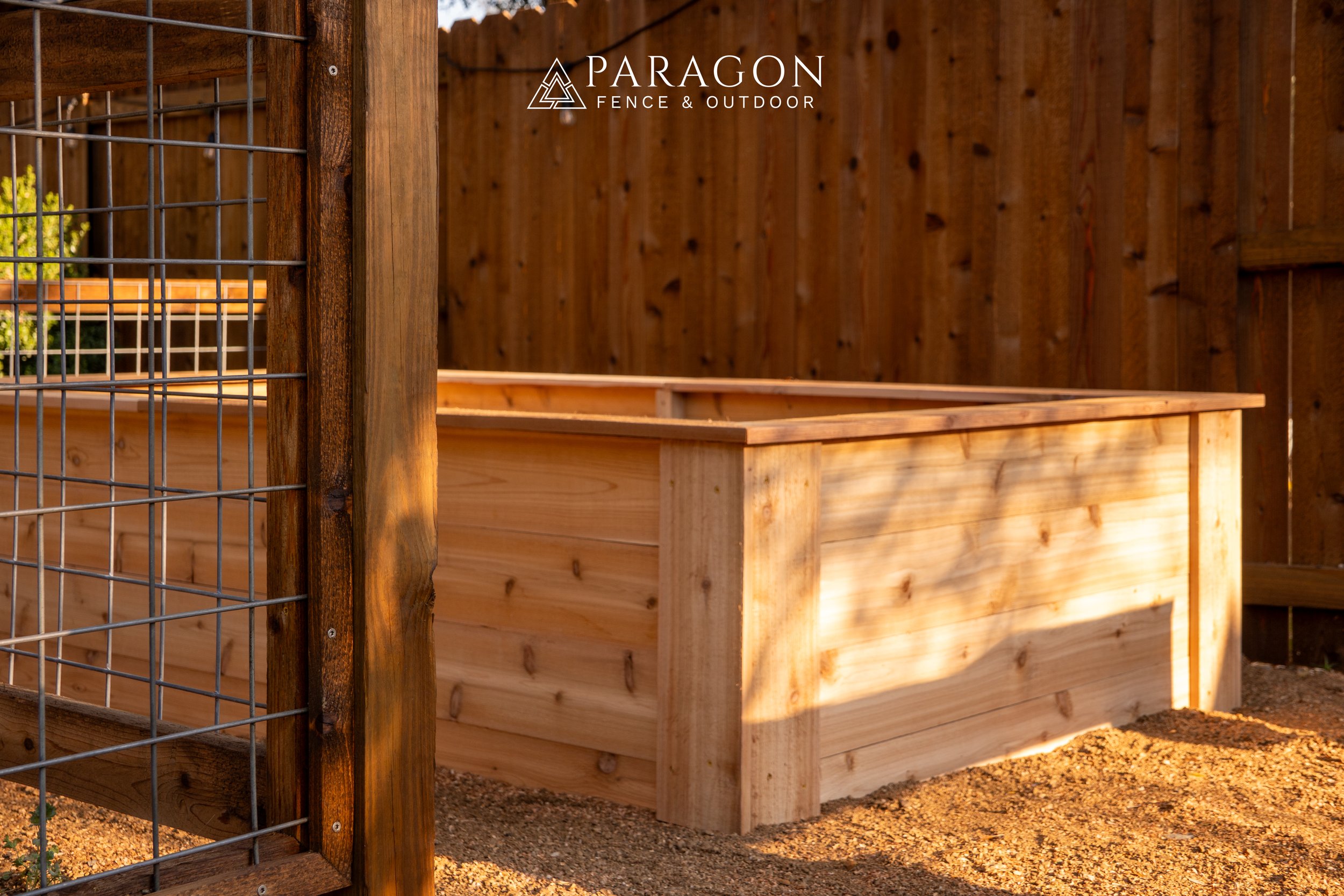 A wooden raised garden bed with fencing in a backyard, with sunlight casting shadows on the soil.