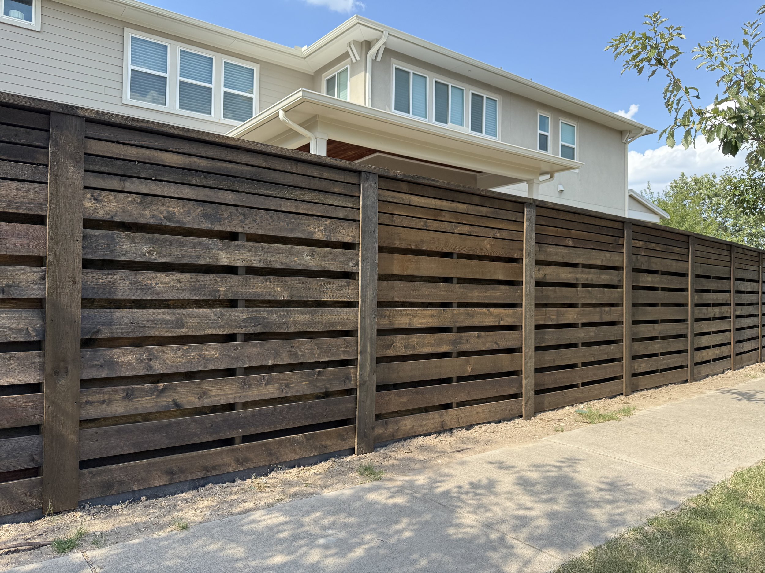 A wooden privacy fence runs along a sidewalk in front of a light-colored, multi-story house with multiple windows and a small porch. Trees are visible in the background under a partly cloudy sky.