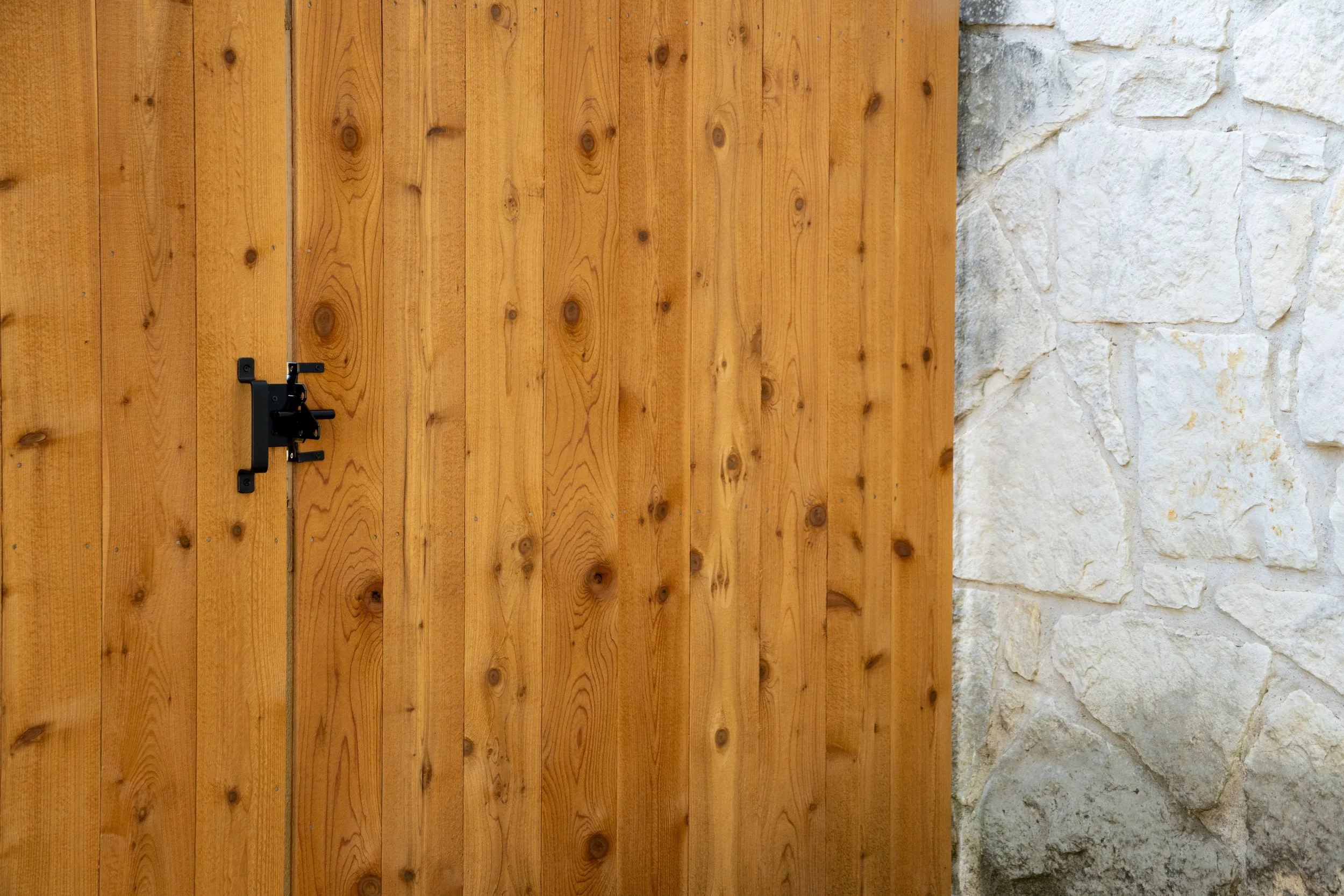 A wooden fence gate with a black latch and hinges, adjacent to a stone wall, with the logo 'Paragon Fence & Outdoor' in the bottom right corner.
