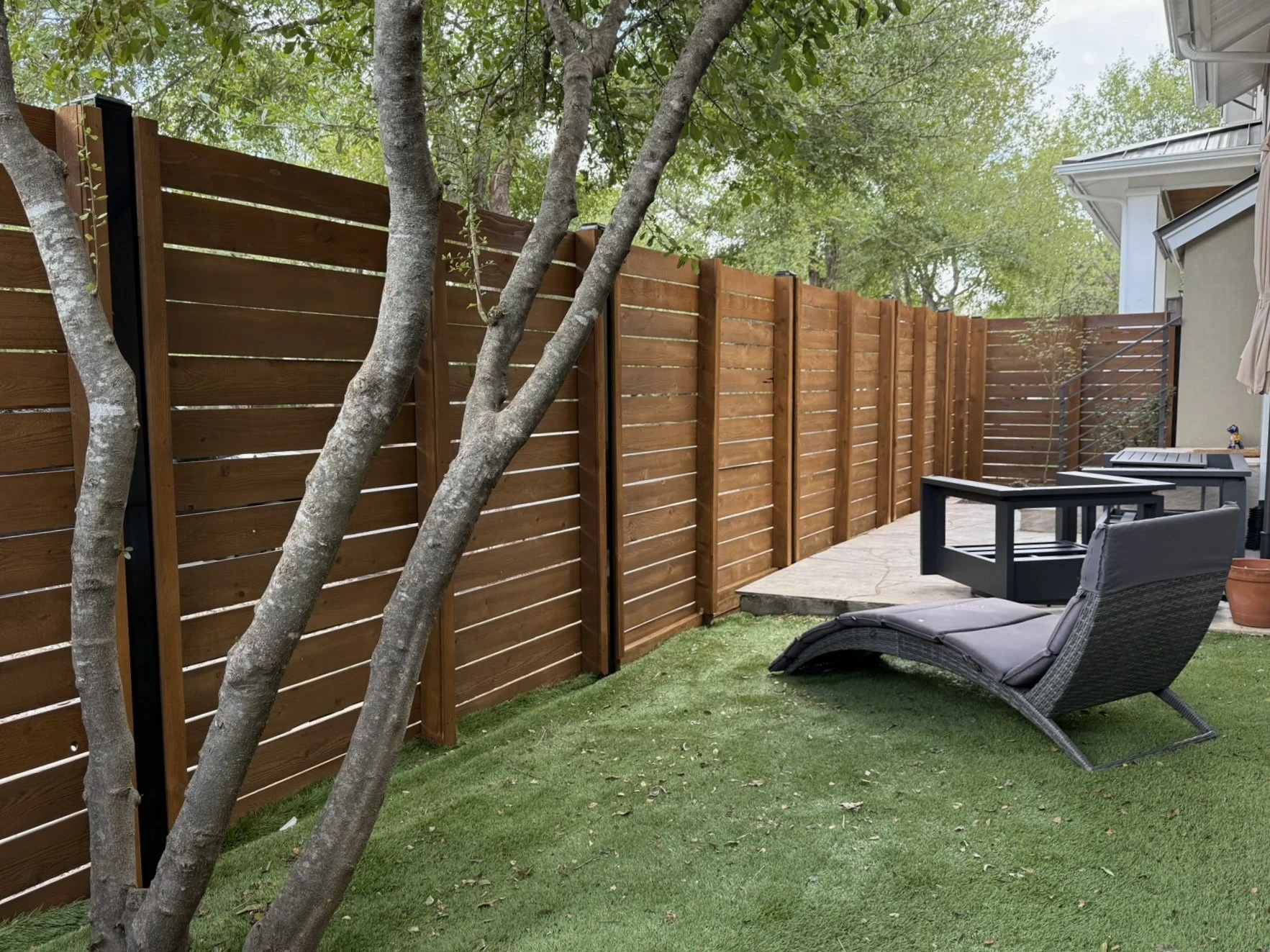 An Austin, TX backyard with a modern, horizontal wooden fence, stained in sable brown with a gray lounge chair on grass, a stone patio, and trees with green leaves.