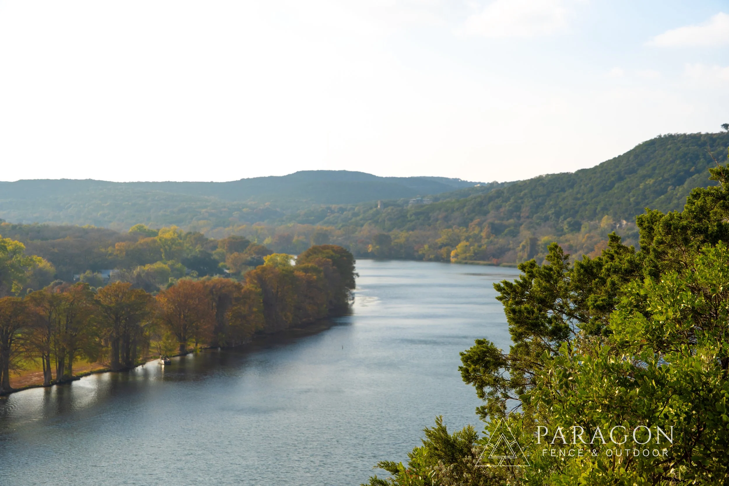 Scenic view of a river winding through lush green and autumn-colored trees with rolling hills in the background, under a partly cloudy sky. Logo of Paragon Fence & Outdoor at the bottom right corner.
