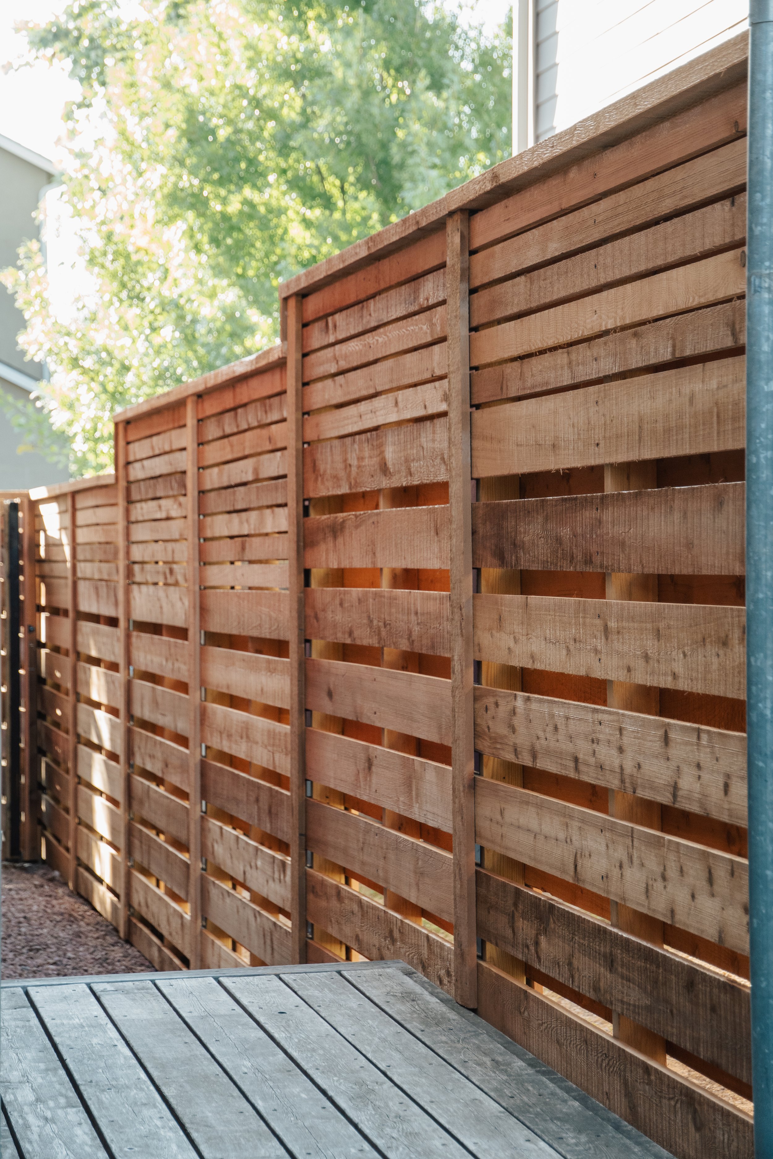 A wooden fence with horizontal slats, installed on a garden bed with soil and some green plants in front of a house with dark siding.