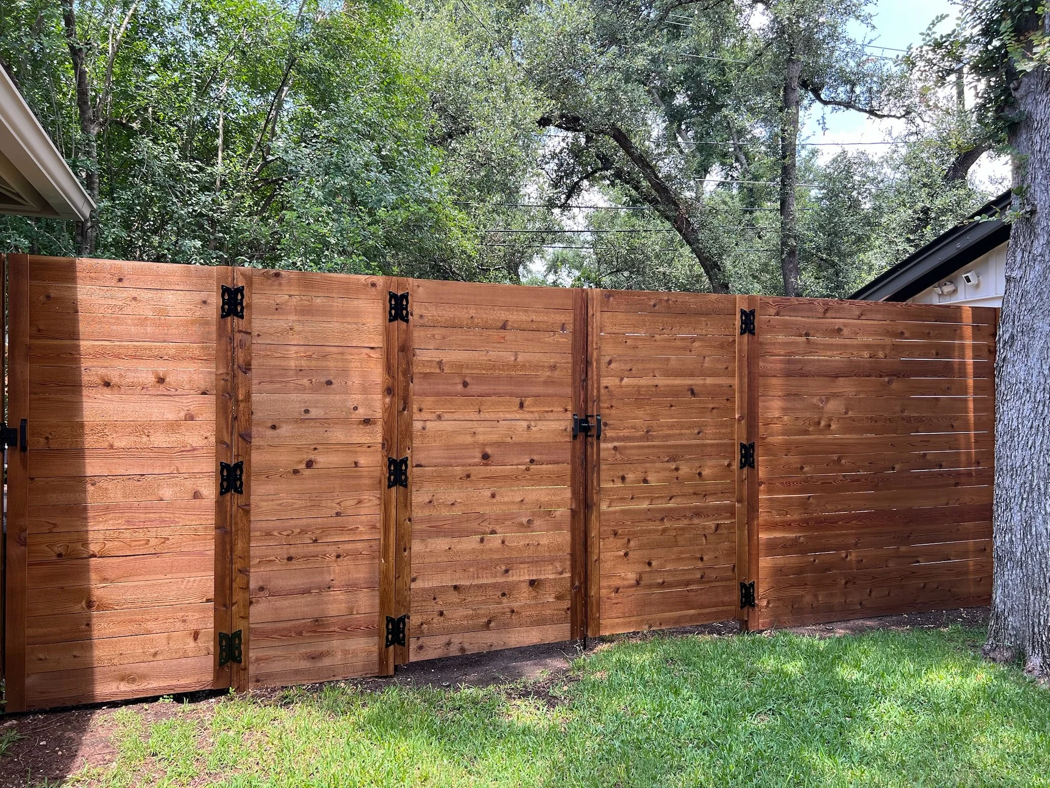 A newly installed wooden privacy fence with black hinges and latch, surrounded by green grass, trees, and part of a house with a sloped roof.