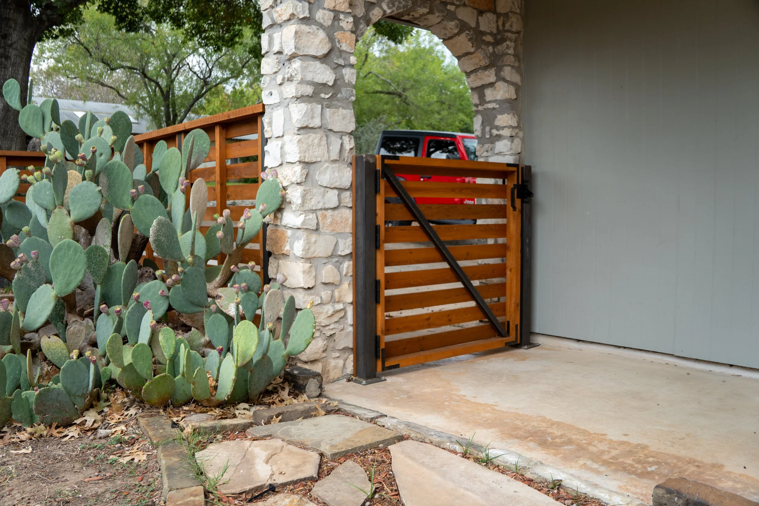 A wooden gate with black hinges is attached to a stone pillar, leading to a driveway where a red vehicle is partially visible. To the left, there are green prickly pear cacti with purple buds. The scene is outdoors with trees and a fence in the background, and the ground features a stone pathway and concrete surface.