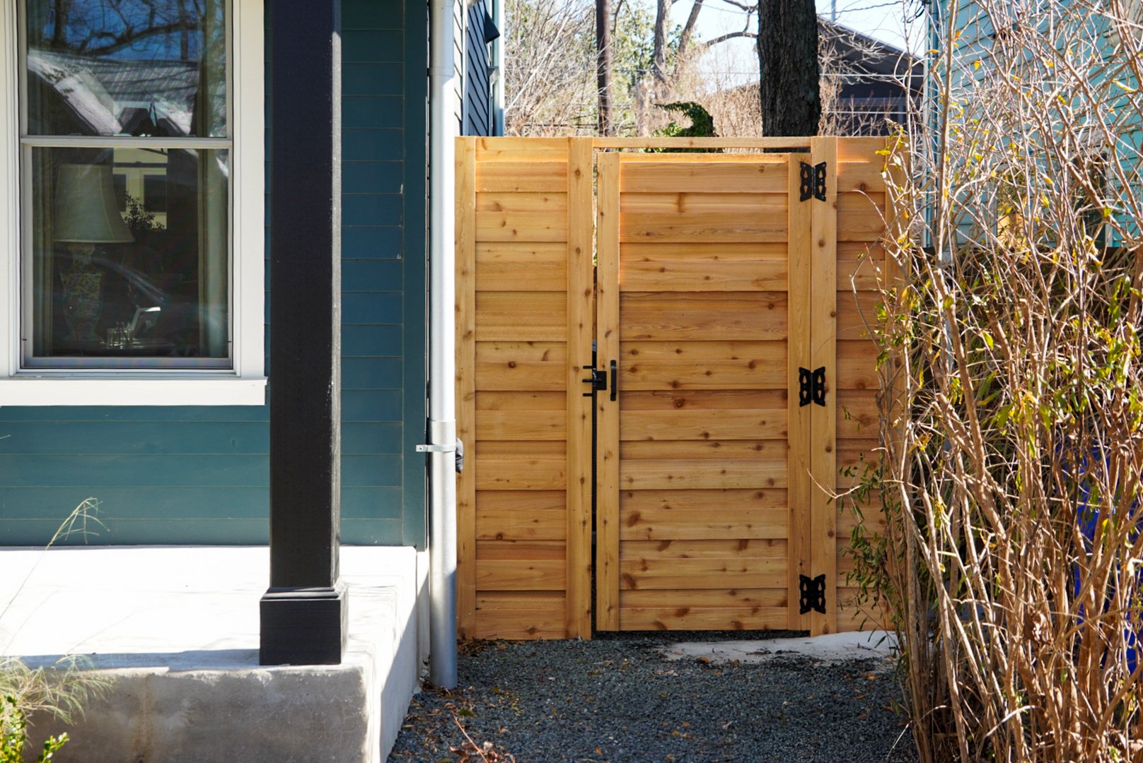 Wooden gate near a house with blue exterior walls, a window, and a black column, surrounded by dry bushes.