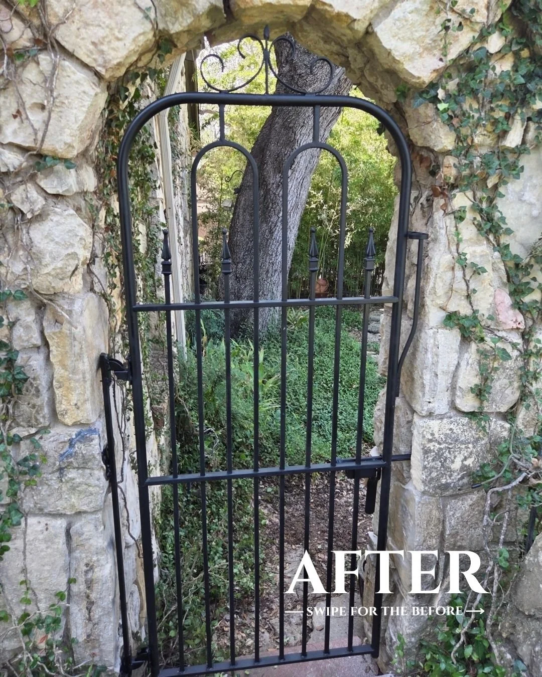 A black metal gate with pointed finials in front of a stone wall, opening to a garden with a large tree and green foliage.