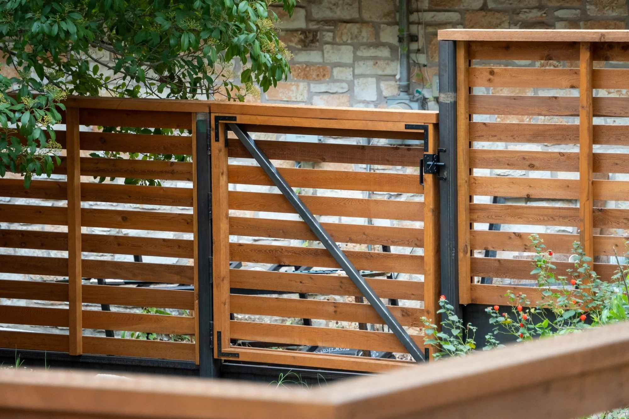 A wooden fence with horizontal slats, installed on a garden bed with soil and some green plants in front of a house with dark siding.