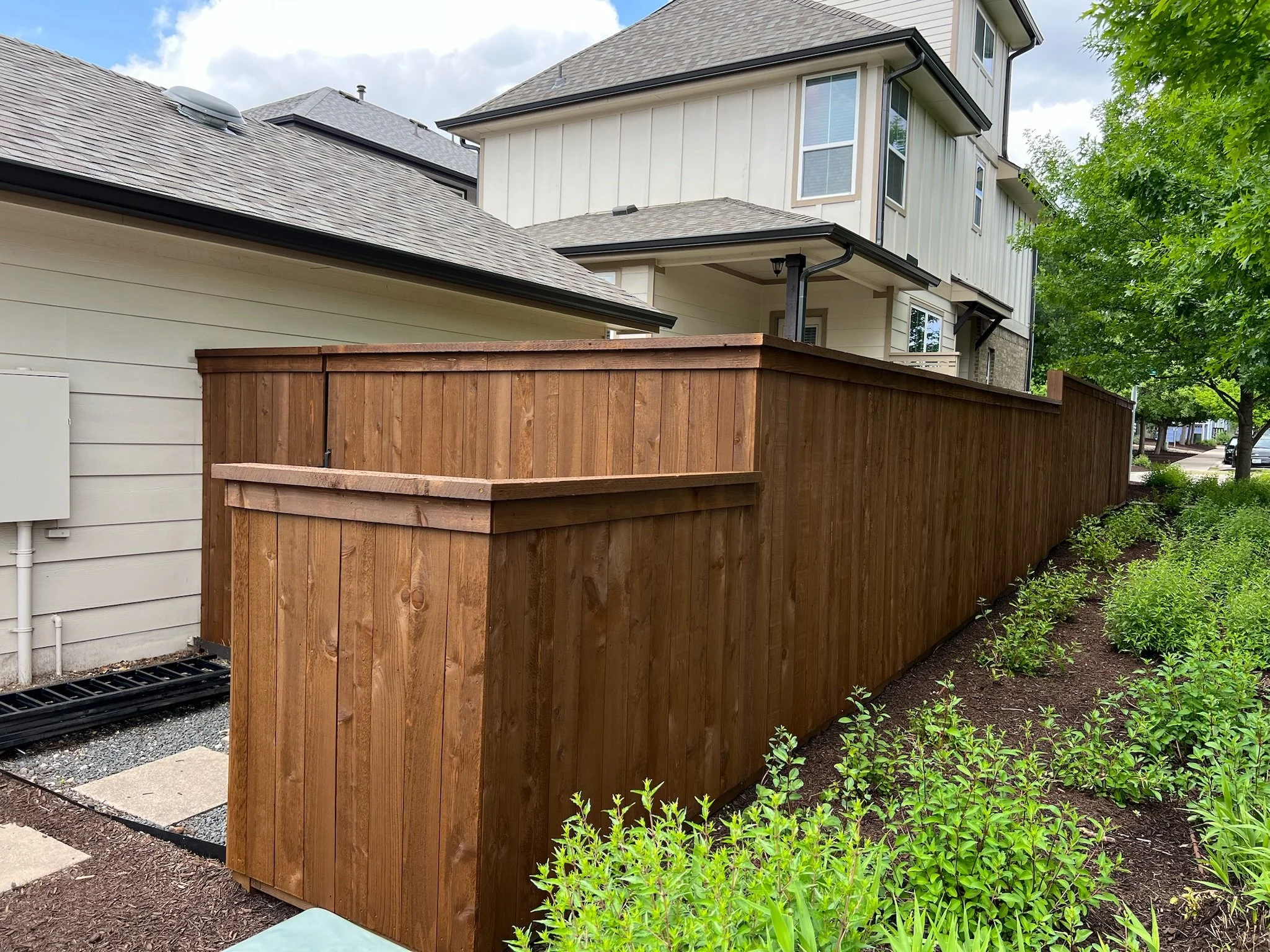 A tall wooden privacy fence surrounds the backyard of a modern house with multiple levels and large windows. There are green bushes and trees along the fence line, and the house has a gray shingled roof.
