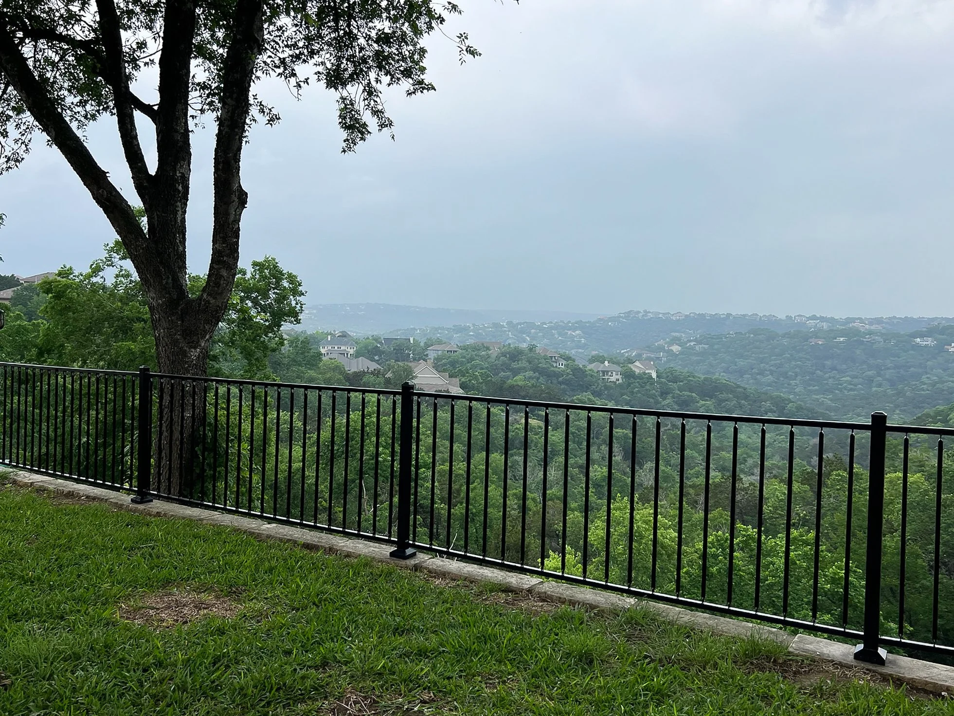 Black metal railing overlooking a lush green valley with trees and distant houses, under an overcast sky.