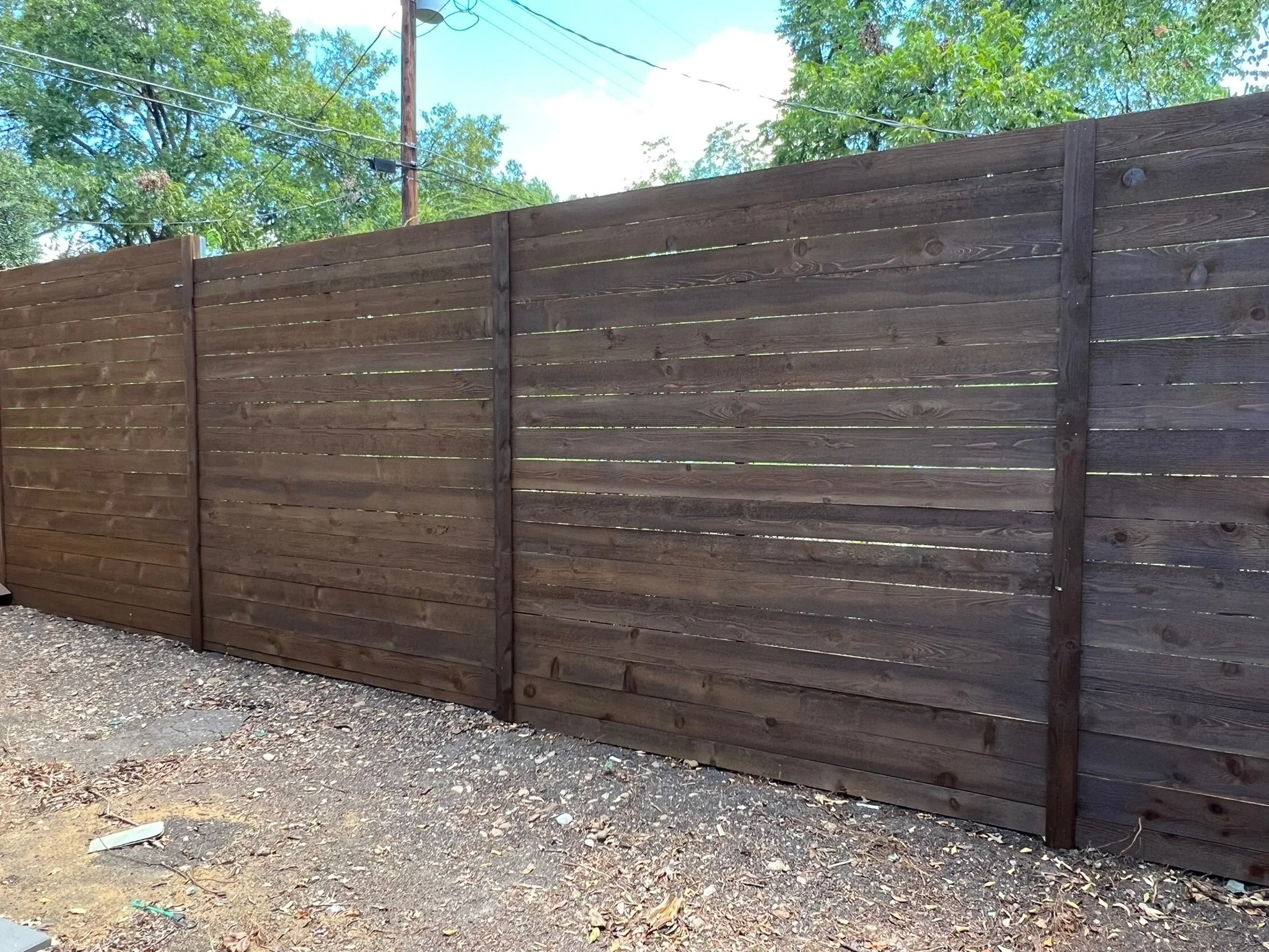 Modern, horizontal wooden privacy fence with burnt hickory stain and vertical support posts, next to a gravel ground, with trees and blue sky in the background.