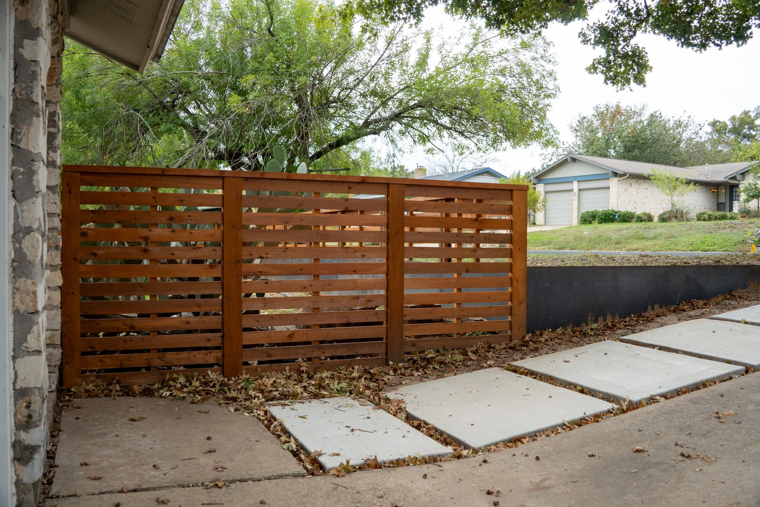 View of front yard with a wooden semi privacy fence, concrete sidewalk, and some fallen leaves. In the background, there is a grassy hill with a house and trees.