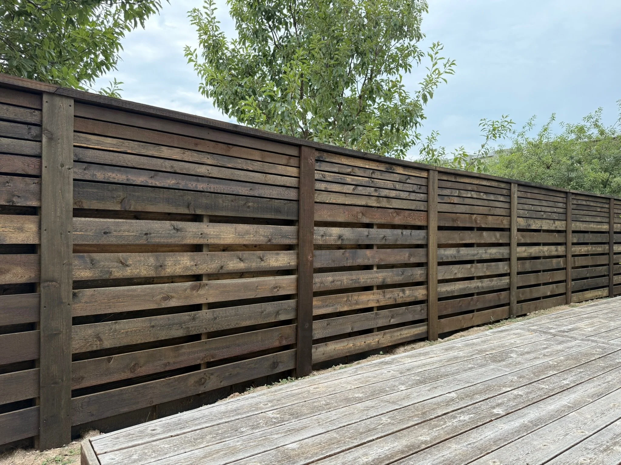 A horizontal wooden privacy fence with burnt hickory stain, and a wooden deck in the foreground. Green trees and a cloudy sky in the background.