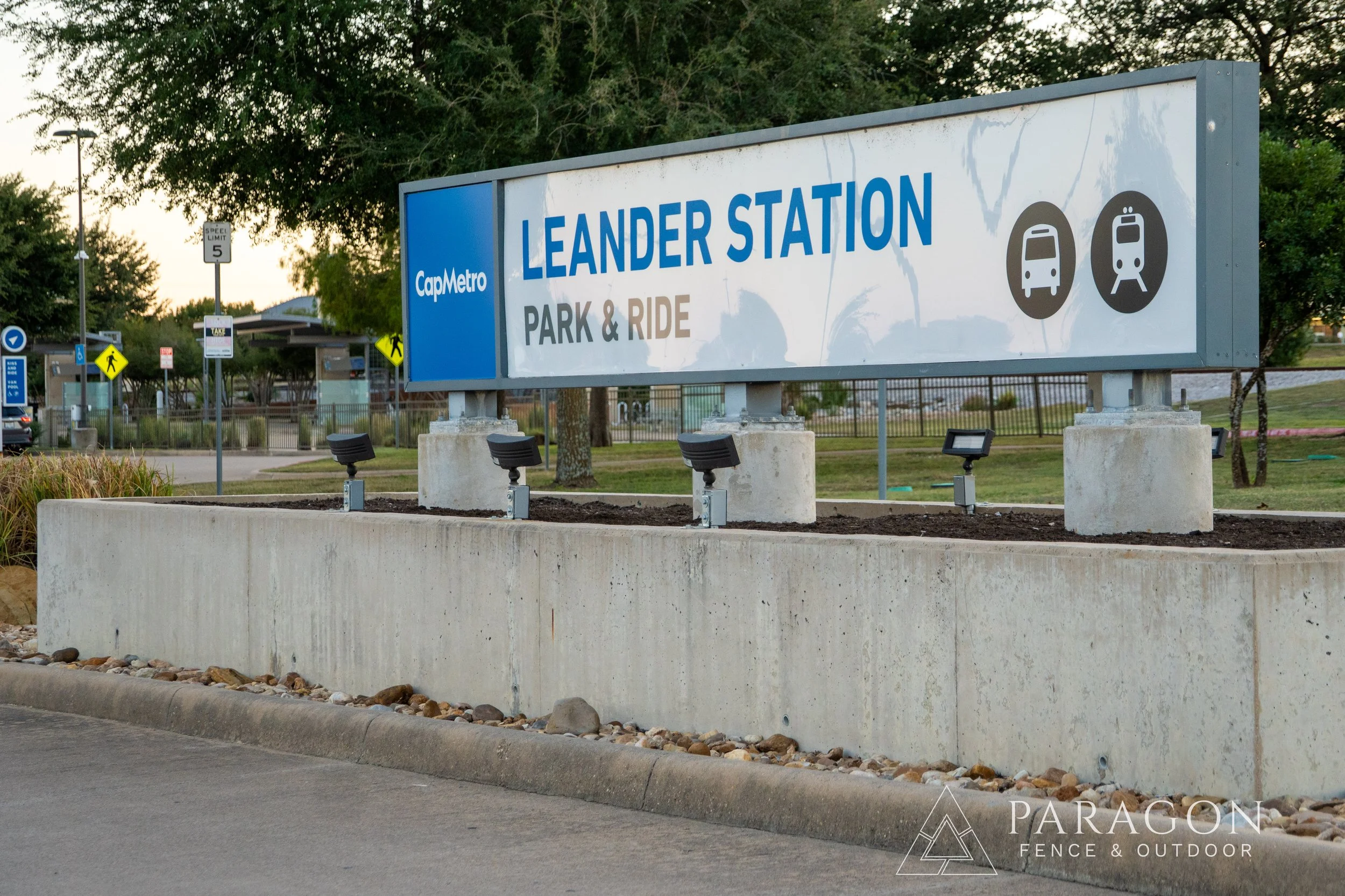 Sign for Leander Station indicating parking and ride, with icons for bus and train, near a parking lot with trees in background.