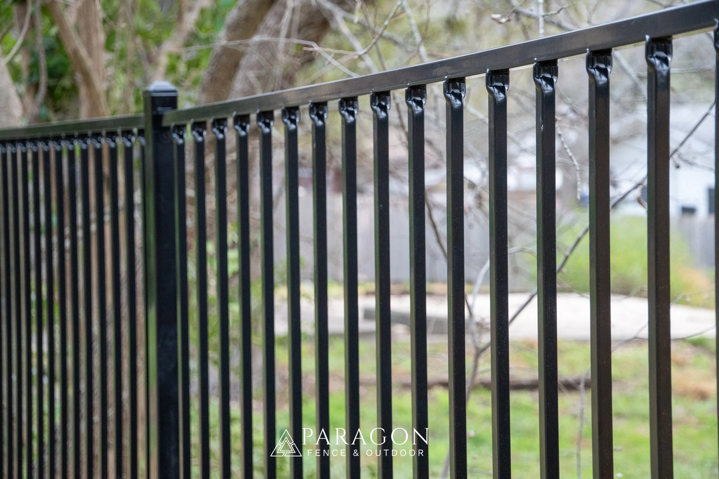 Black metal fence with vertical bars and decorative tips in an outdoor setting with trees and grass in the background.