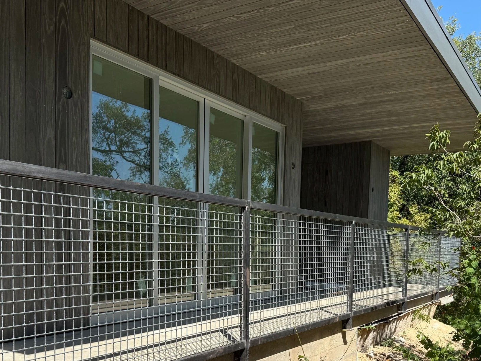 A modern house balcony with wood paneling and large glass sliding windows, surrounded by green trees.