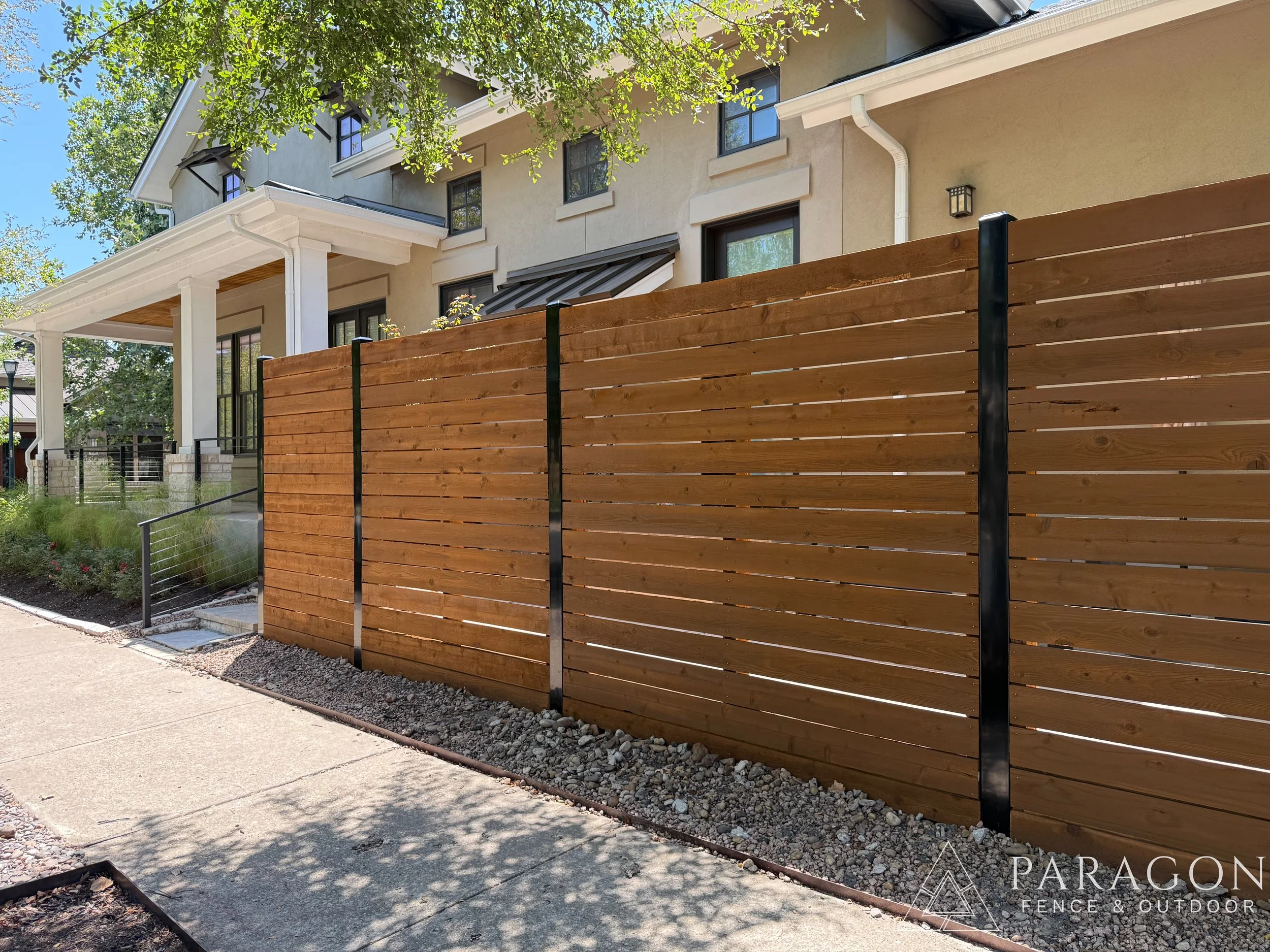 New wooden fence installed in front of a beige residential building with large windows and white pillars. The fence is supported by black metal posts and is along a concrete sidewalk with gravel underneath.