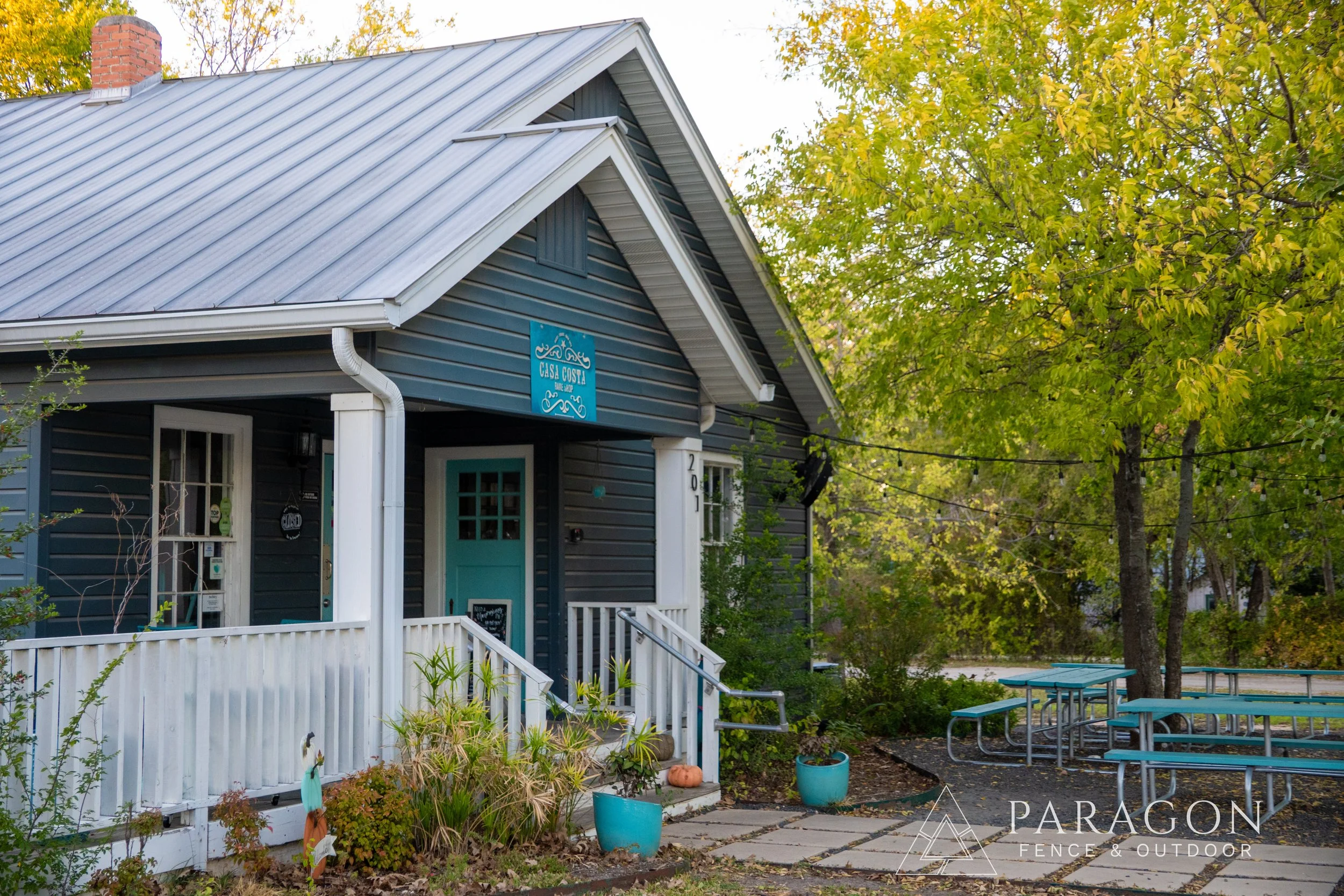 A blue house with white trim and a metal roof, decorated with a sign that reads "Casa Costa." There are potted plants and pumpkins near the front porch, with trees in the background and outdoor picnic tables on the right.