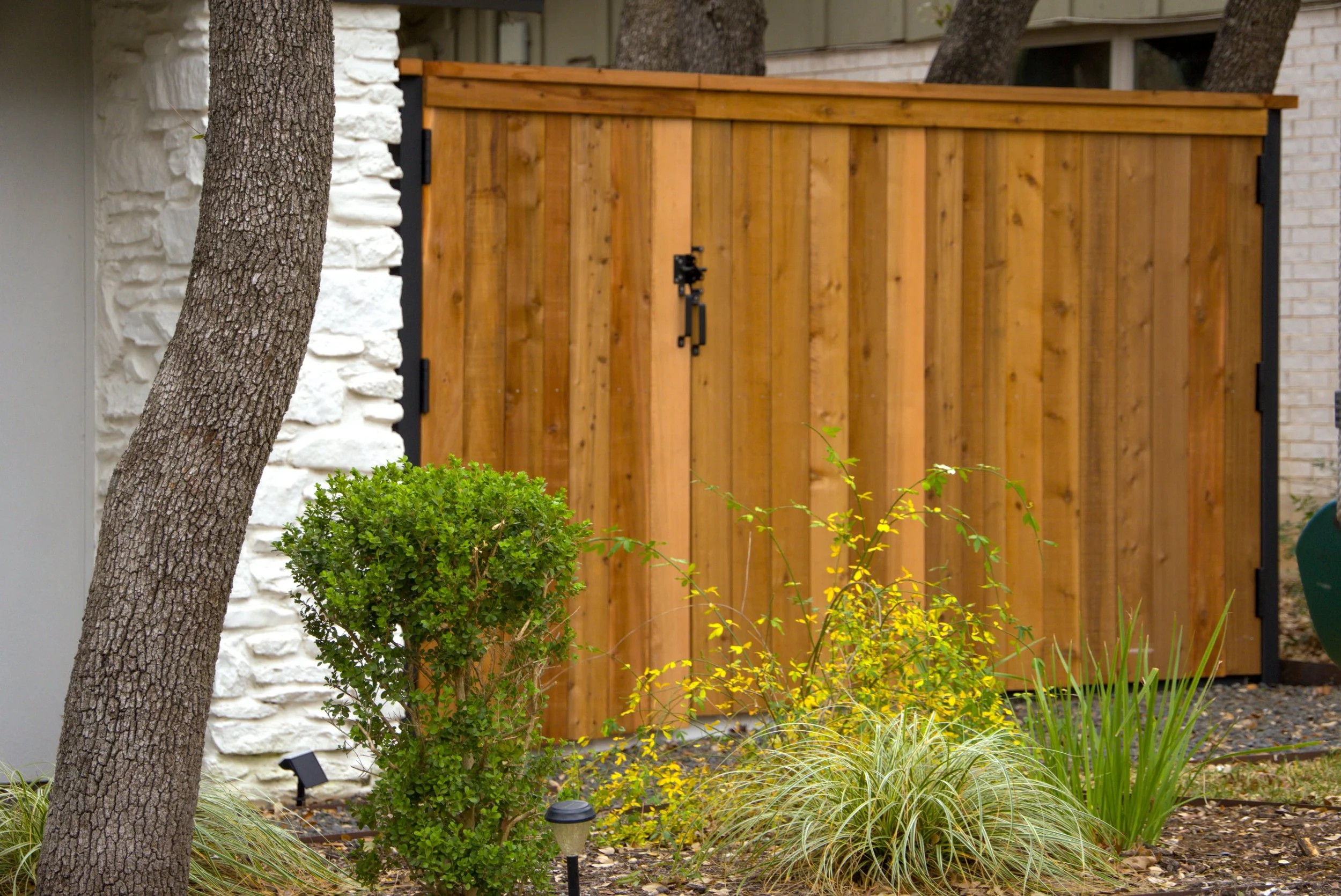 The image shows the side of a house with a gray metal roof, a black-framed window, and a wooden gate made of vertical planks attached to a white brick wall. There are trees and grass in the background and some plants in the foreground.