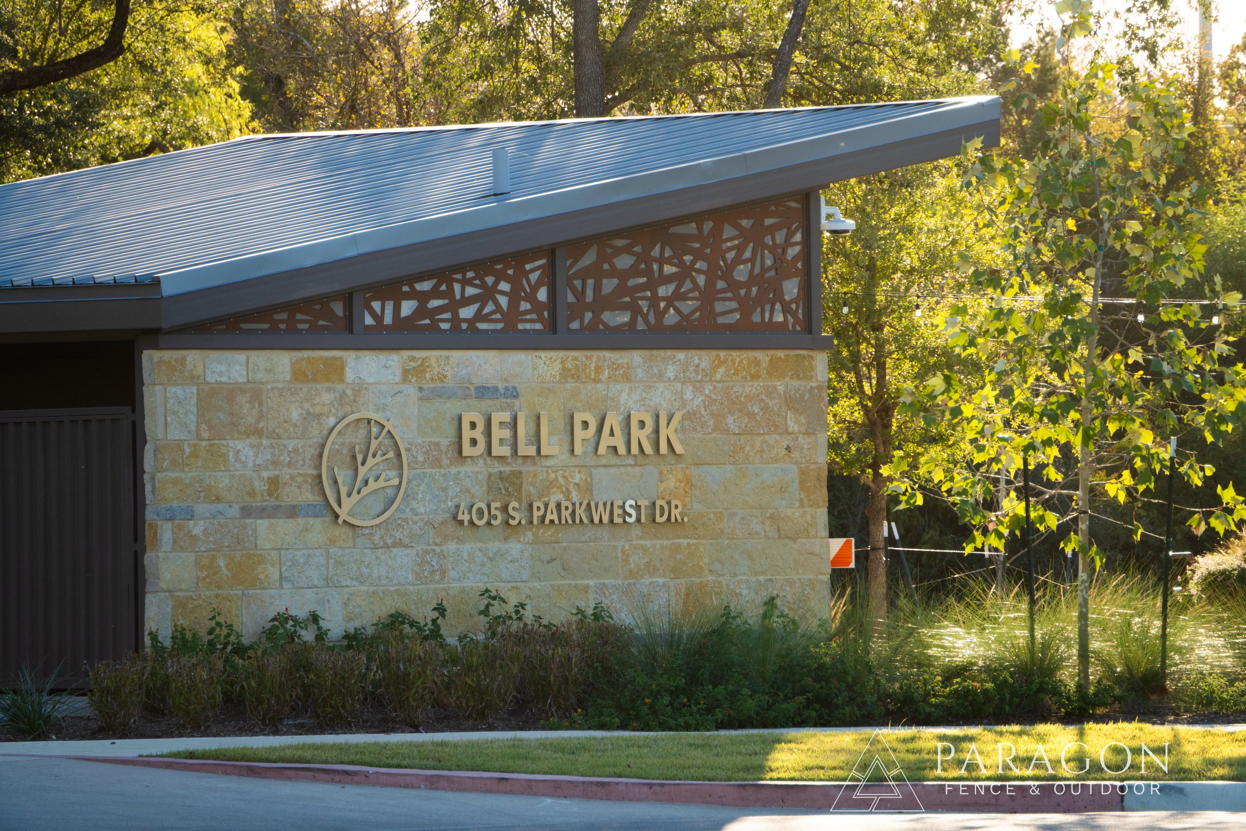 Entrance sign for Bell Park, located at 405 South Park West Drive, with a stone wall, a metal logo of a tree, and surrounded by trees and greenery, during daylight.