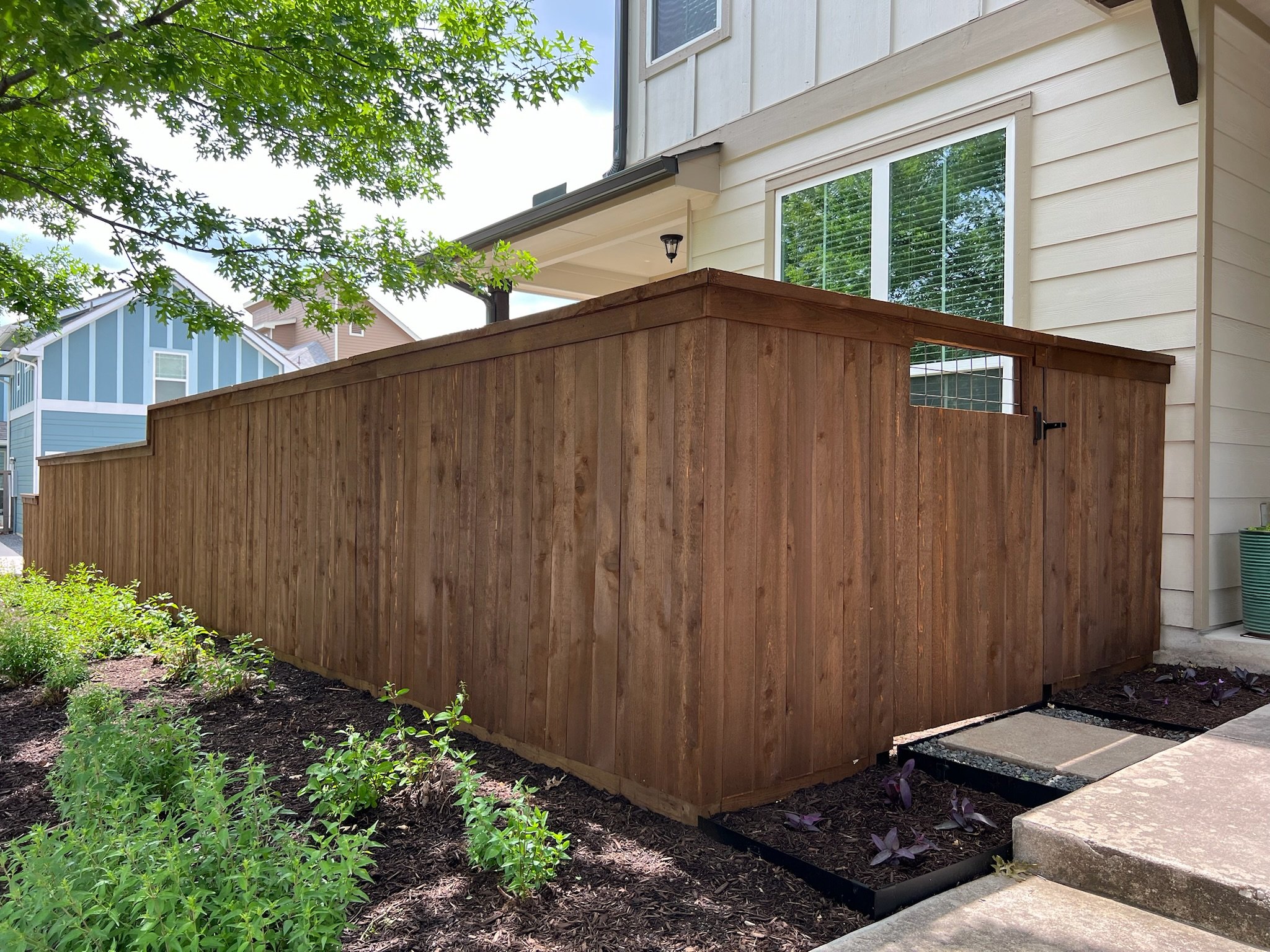 Newly constructed vertical wooden privacy fence stained in coffee brown with a latch gate adjacent to a beige house with large windows, surrounded by garden plants and mulch, under a partly cloudy sky.