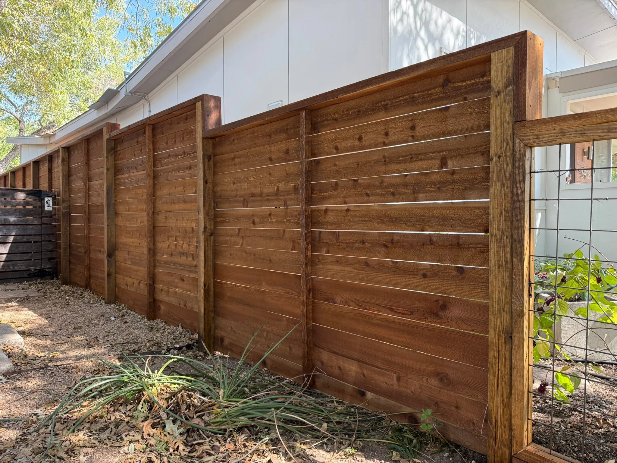 Horizontal wooden privacy fence, adjacent to a white home with a window, and some plants and rocks in the garden.