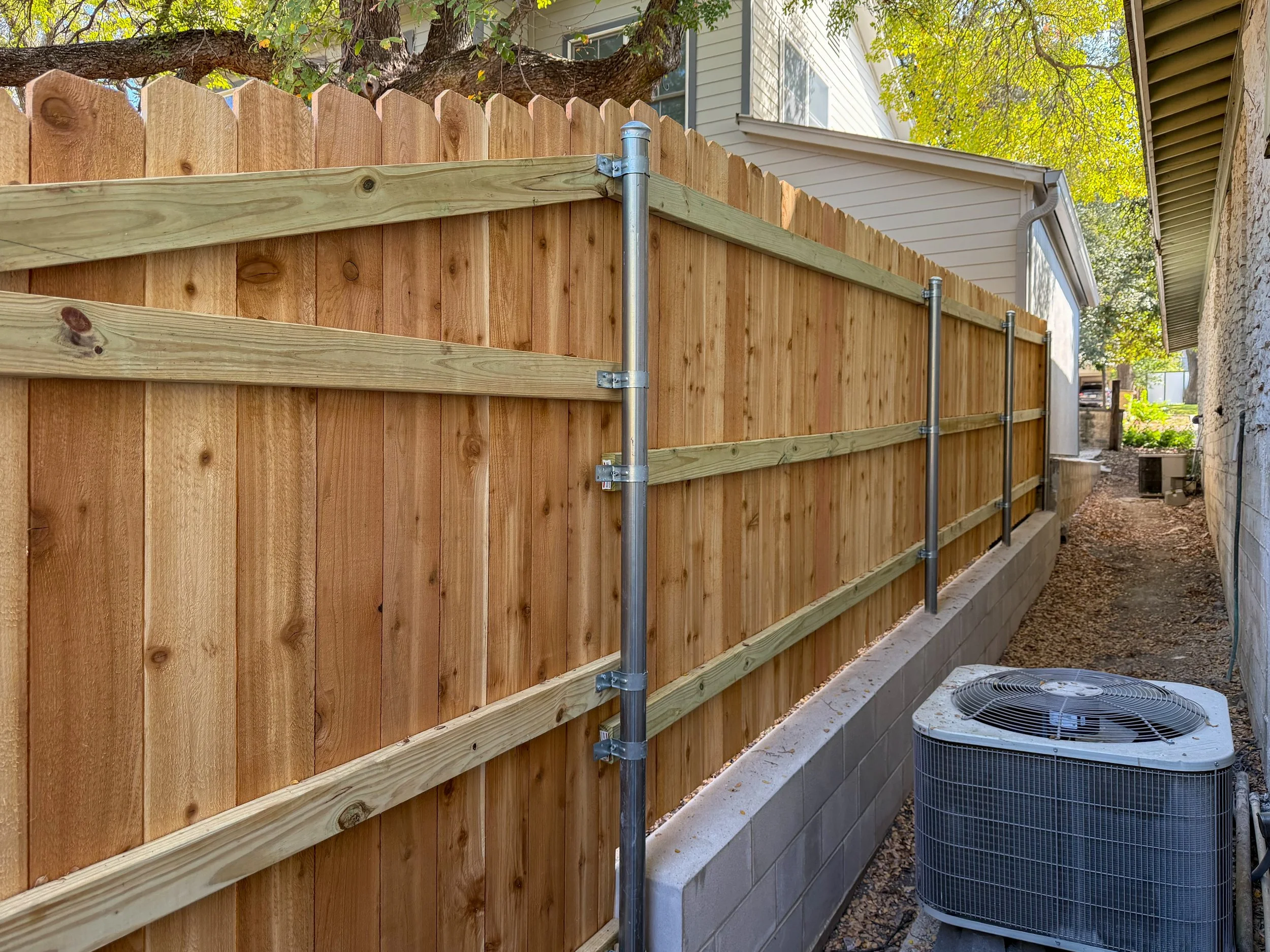 Side view of a newly installed wooden privacy fence with metal support poles, next to a house with an air conditioning unit on the ground.