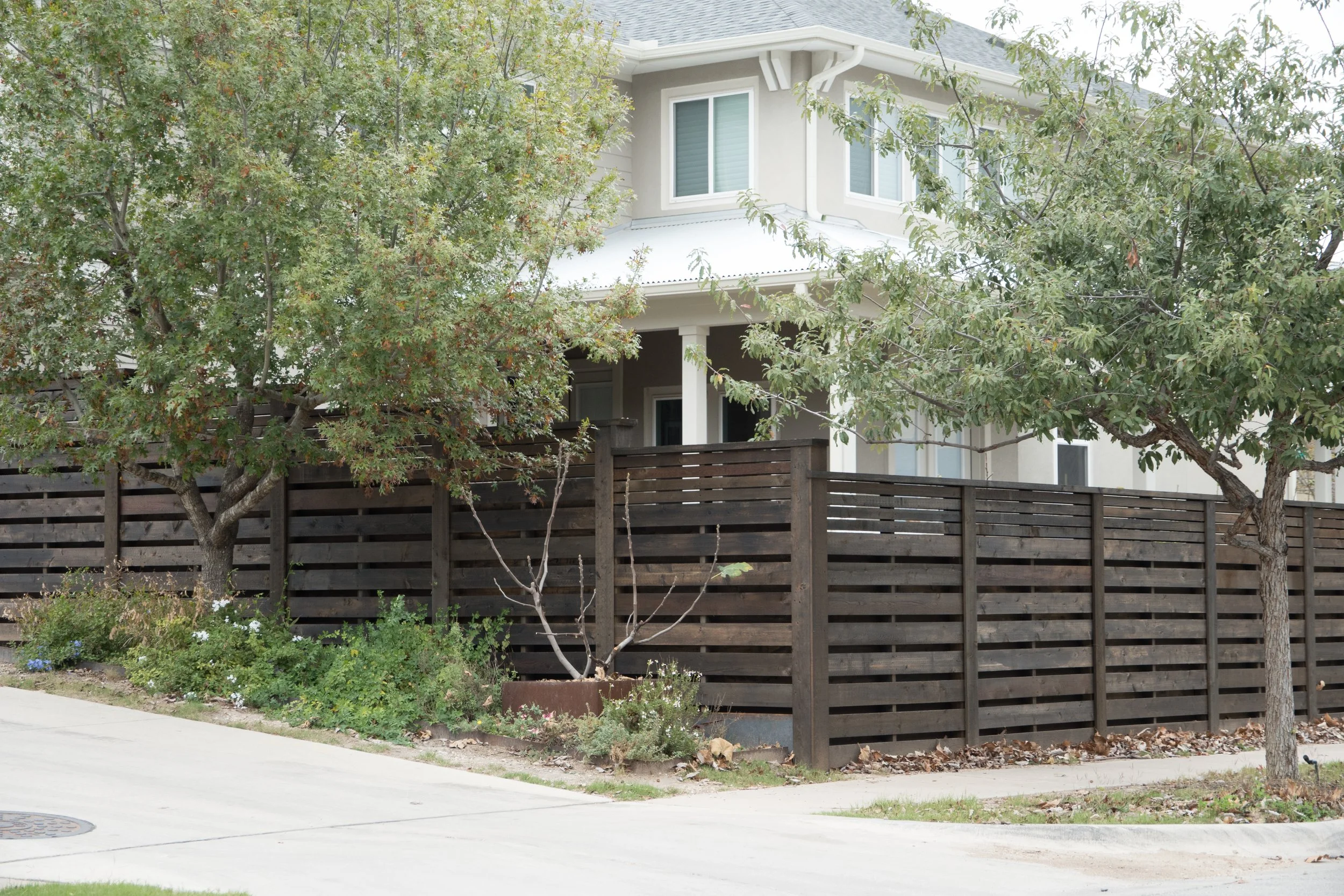 A wooden horizontal shadow box privacy fence surrounds a residential backyard, with trees and plants in front of it, and a house visible behind the fence.