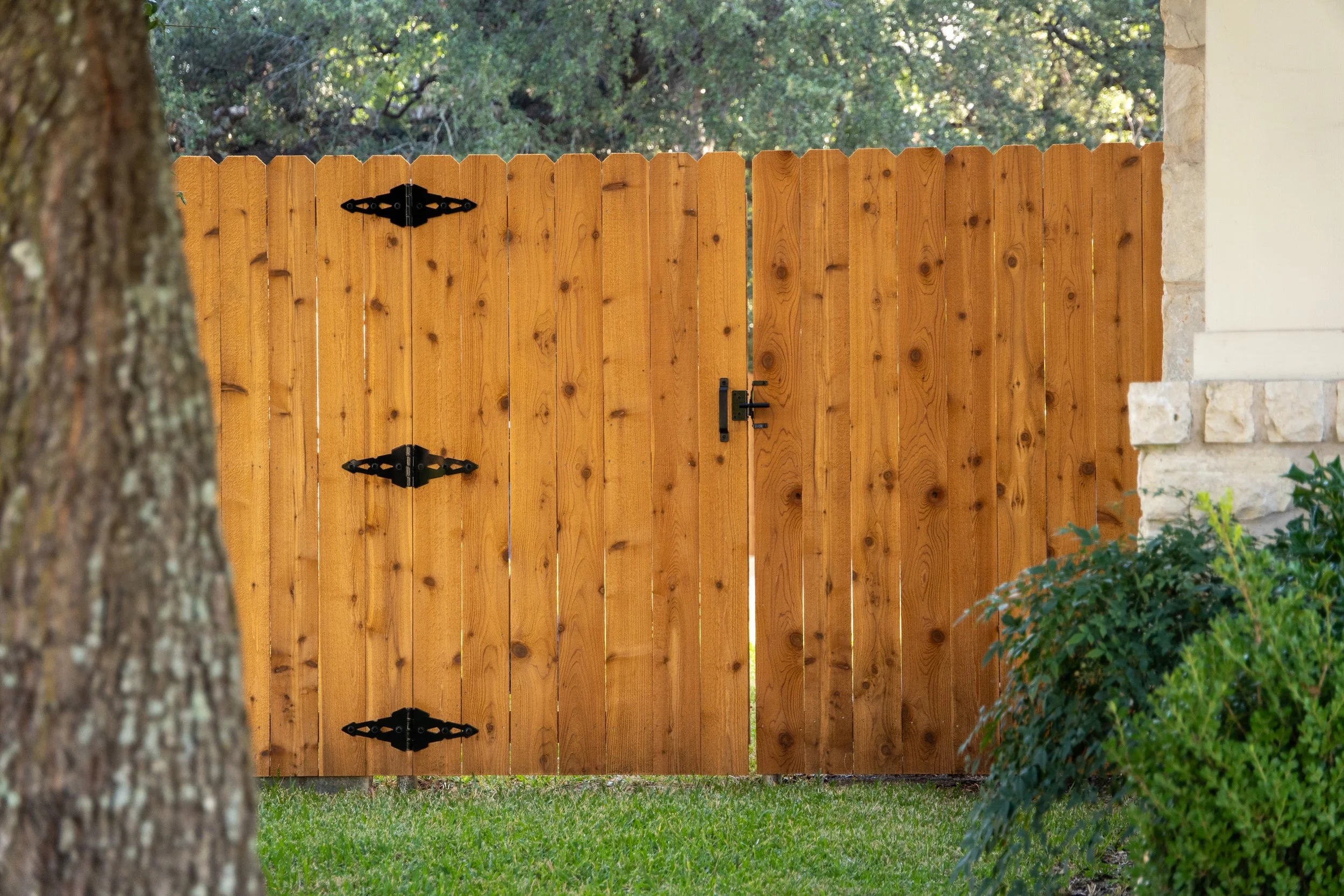Wooden cedar fence with black hinges and latch, partially covering a stone wall and surrounded by green shrubs and grass.