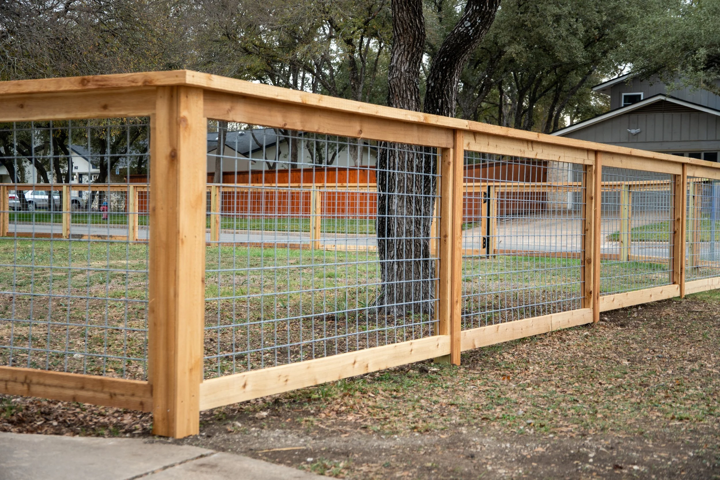 A wooden and wire mesh fence installed in a yard with trees and houses visible in the background, with a logo that says 'Paragon Fence & Outdoor' in the bottom right corner.