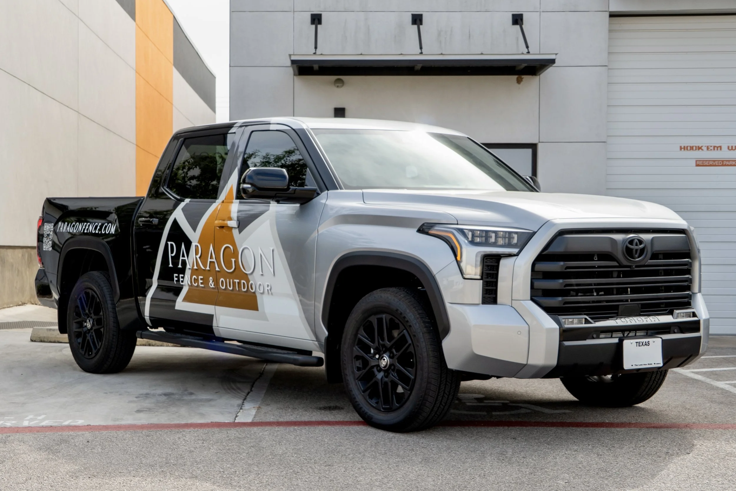 A silver and black Toyota pickup truck with a logo and website for Paragon Fence & Outdoor on its side, parked in an outdoor lot near a modern building.