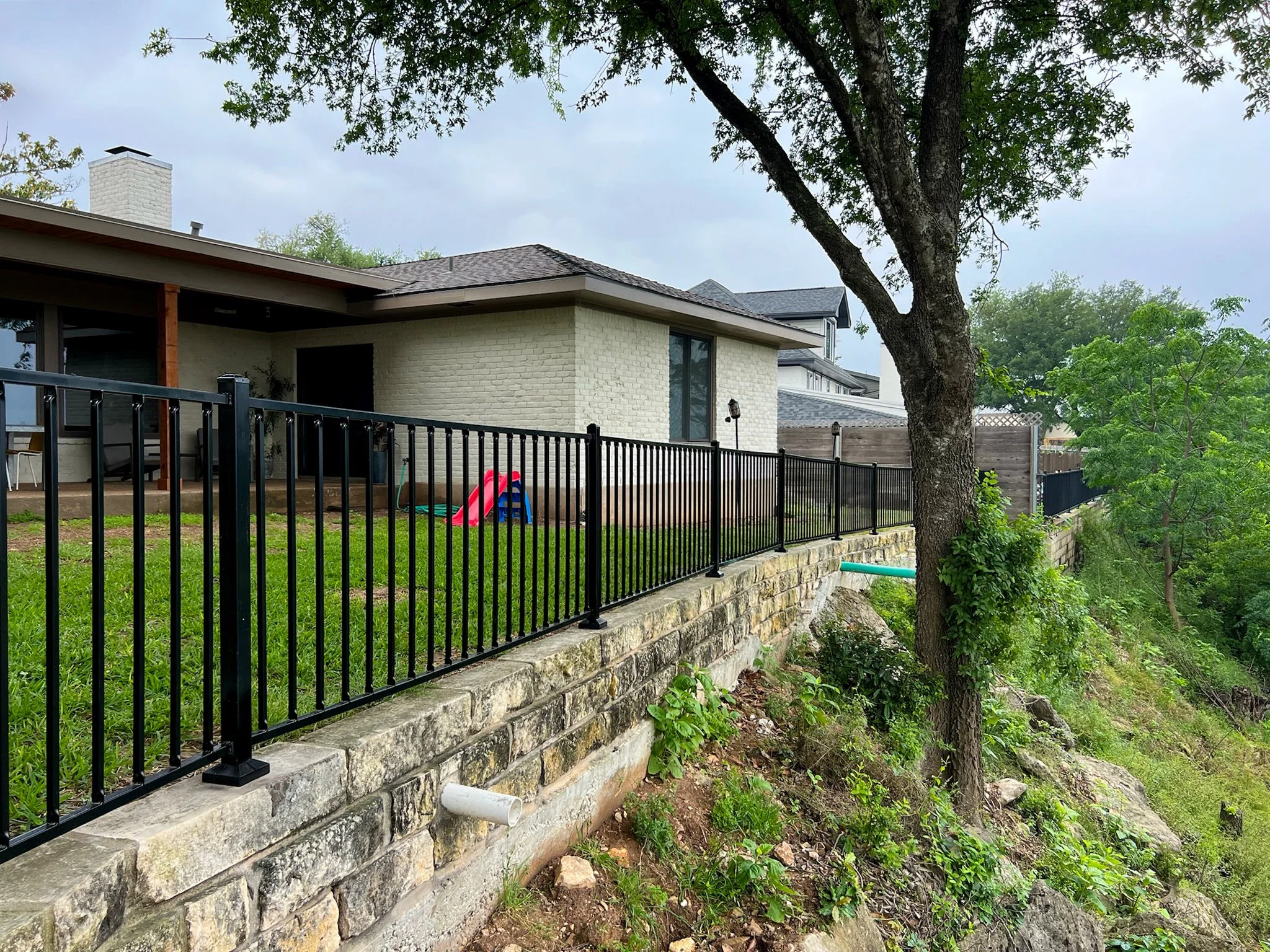 Black metal fence in a wooded outdoor area with green trees and shrubs.