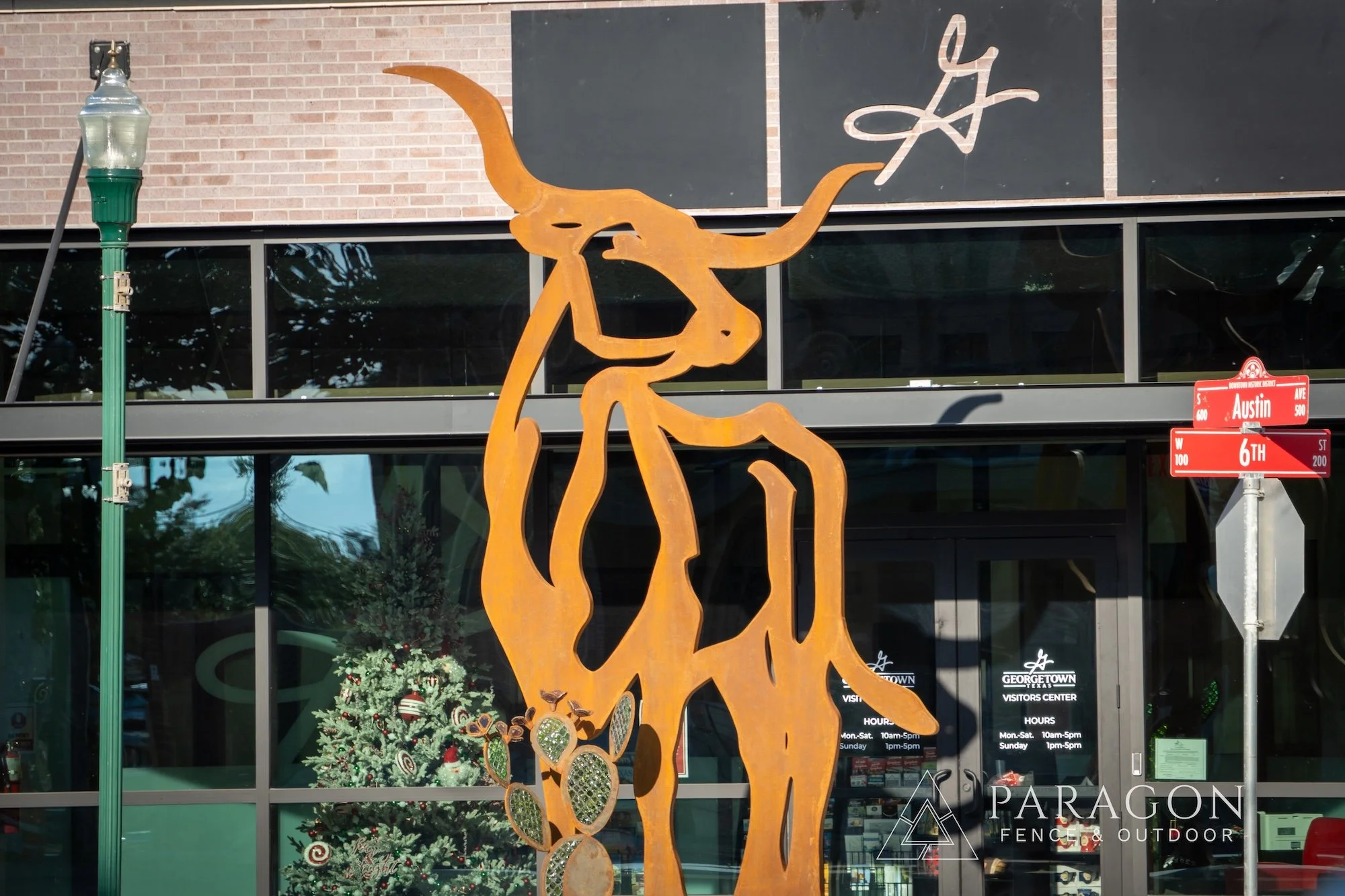 Rust-colored metal sculpture of a giraffe with a long neck, horns, and a tail, displayed in front of the Georgetown visitors center, with Christmas decorations visible inside.