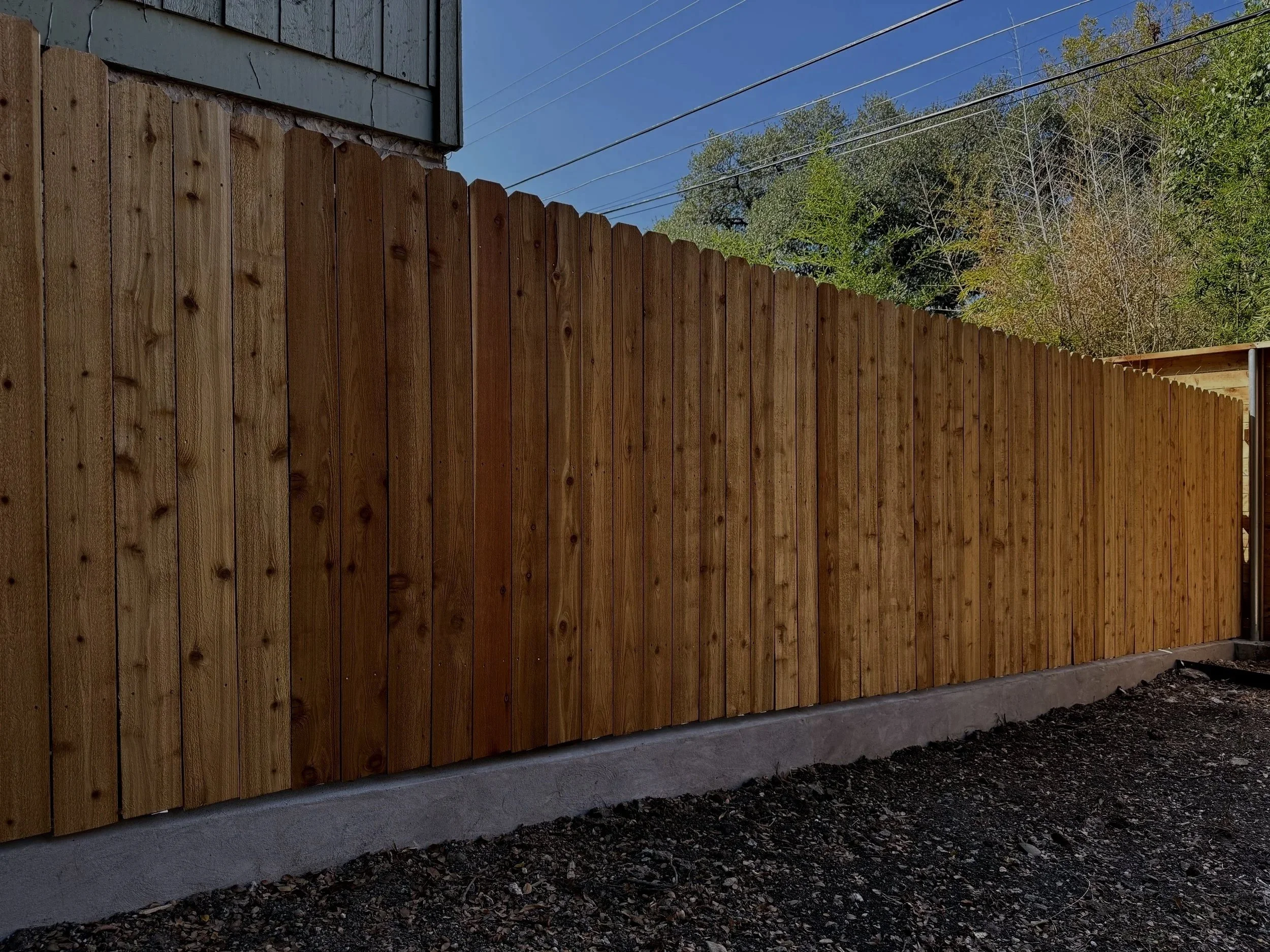 New wooden privacy fence installed on a concrete foundation, with a gravel path in front, against a background of trees and a blue sky.