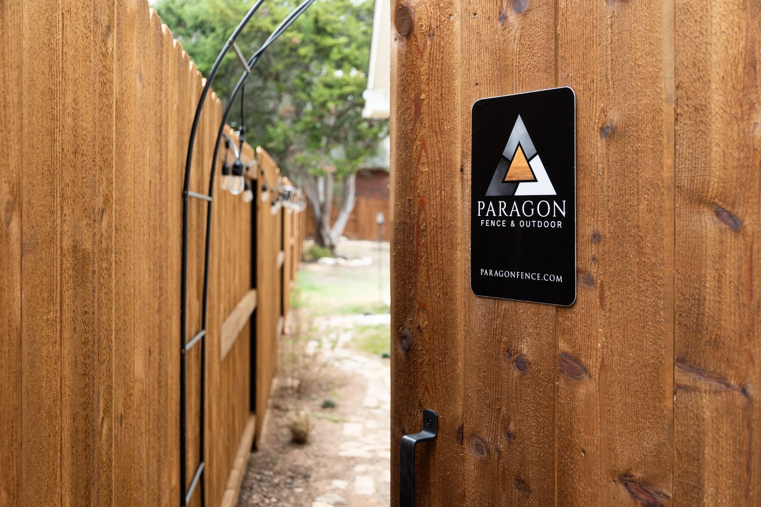 Close-up view of a wooden fence with a black sign that reads 'Paragon Fence & Outdoor' and the website 'paragonfence.com'. The fence is lined with string lights, and a path is visible in the background.