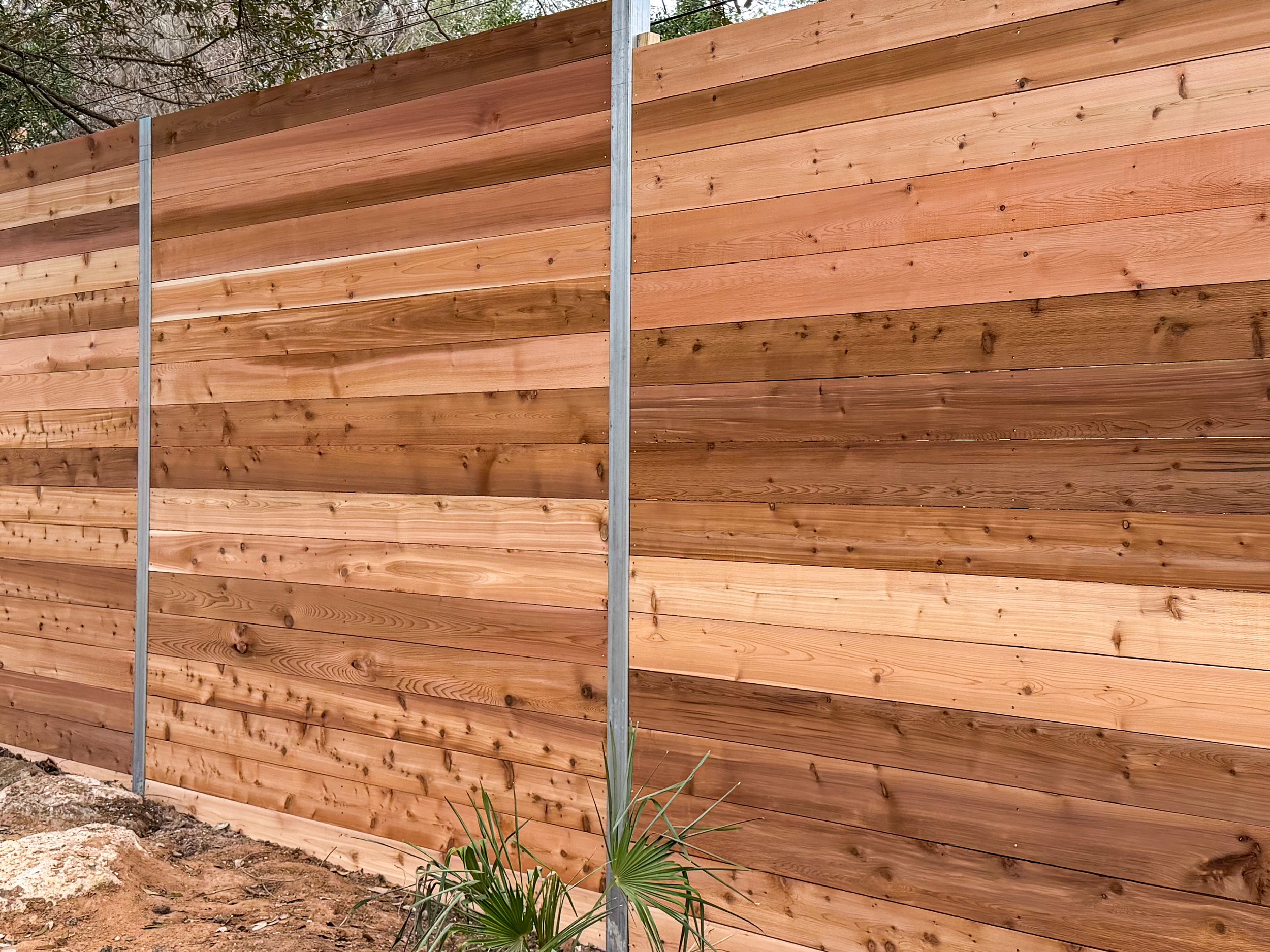 A wooden privacy fence made of horizontal planks with metal posts at intervals, with some plants and rocks at the base.