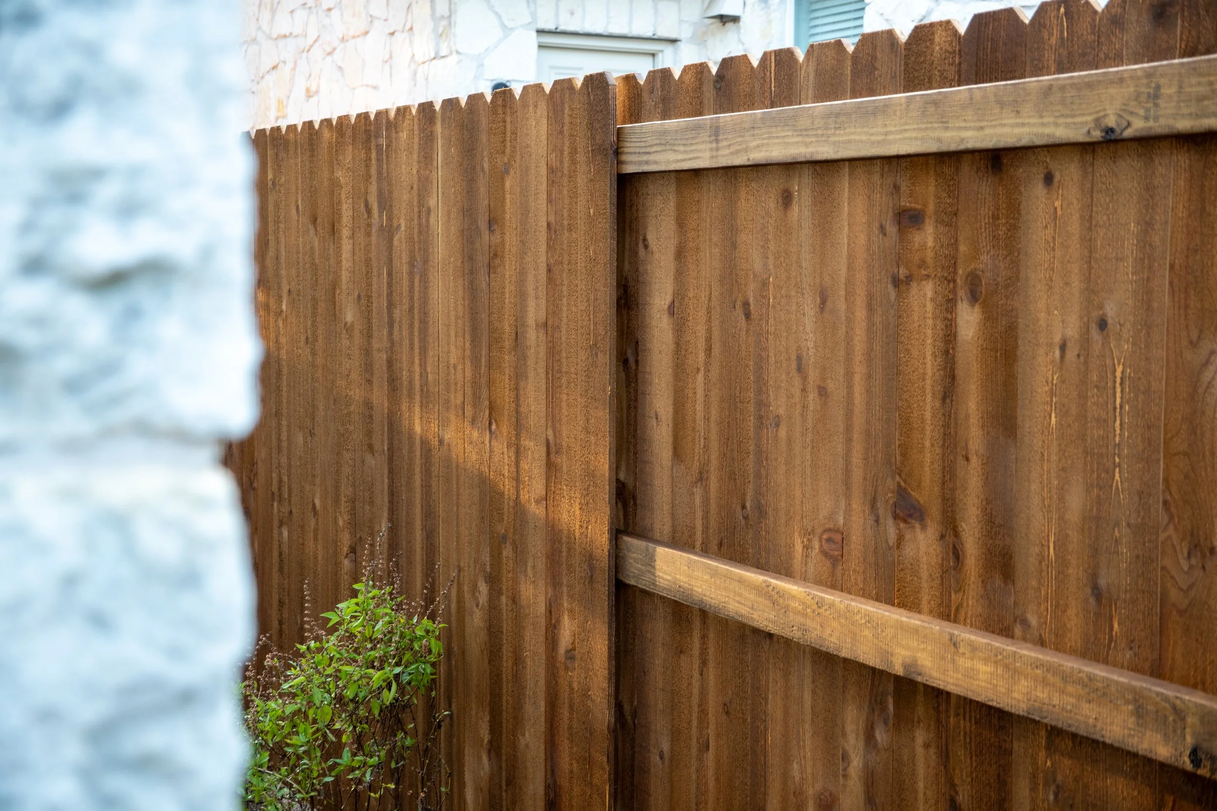 Wooden privacy fence with a small green bush at the base, partially obscured by a blurred white wall in the foreground.