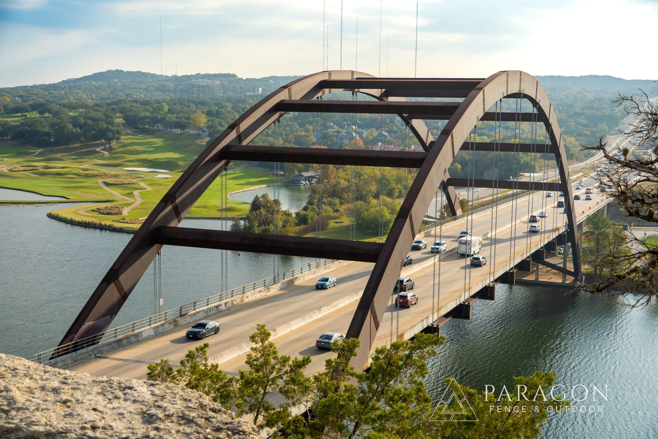 A large steel arch bridge over a river with cars traveling on it, green landscape and golf course visible in the background.