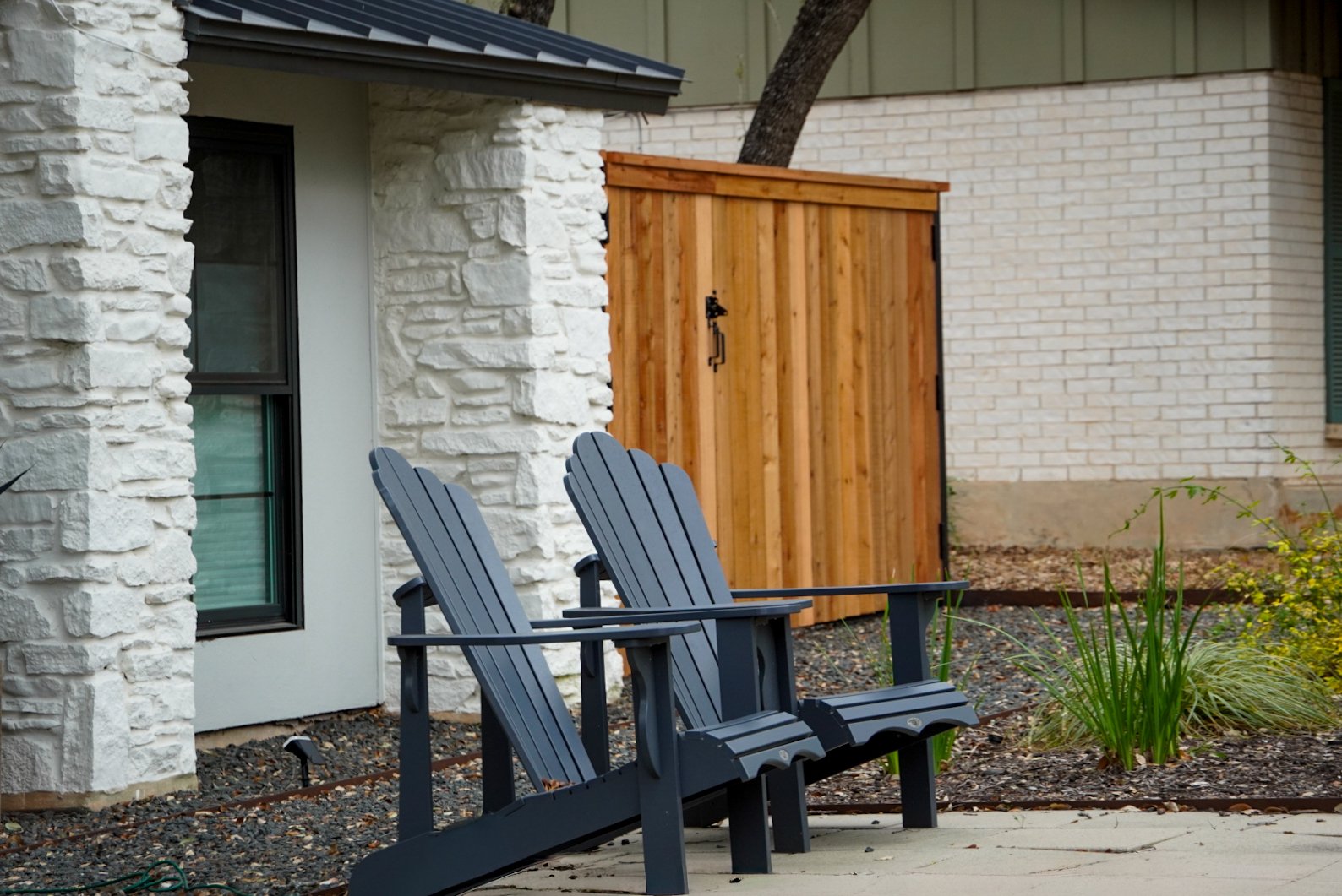 Two gray Adirondack chairs on a concrete patio in front of a white brick house with a black window and a small garden bed with green plants. A wooden storage shed is in the background.