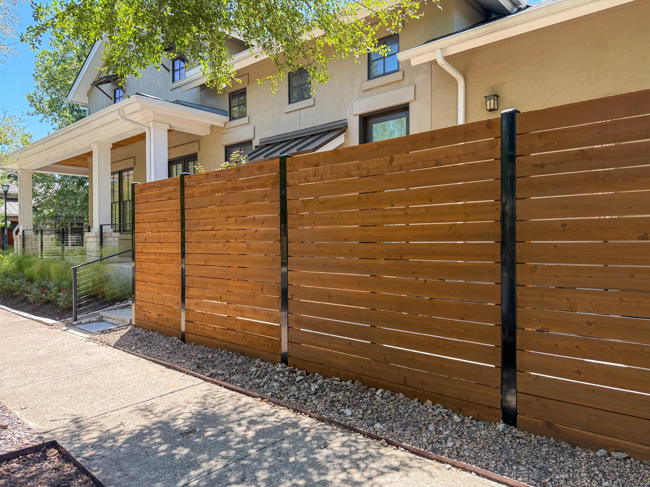 A wooden privacy fence with horizontal slats and black metal support posts in front of a residential house. The house features a porch, large windows, and a beige exterior with dark framing. There is a sidewalk in the foreground, with some small rocks along the base of the fence and trees providing shade. The scene is bright and sunny.