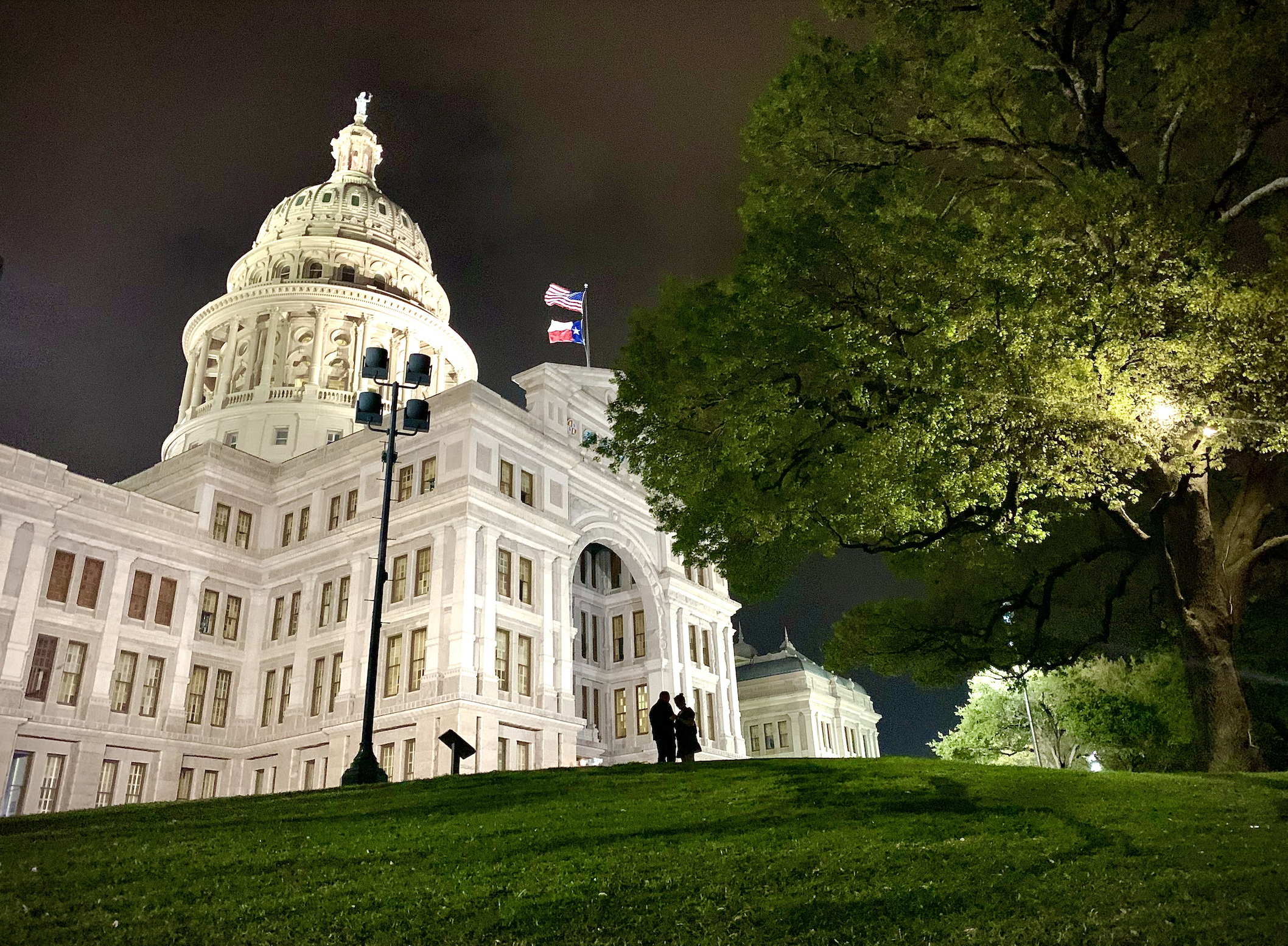 Night view of the Texas State Capitol building in Austin, with two people standing on the grass near the entrance, illuminated by exterior lights, and a large leafy tree on the right side.