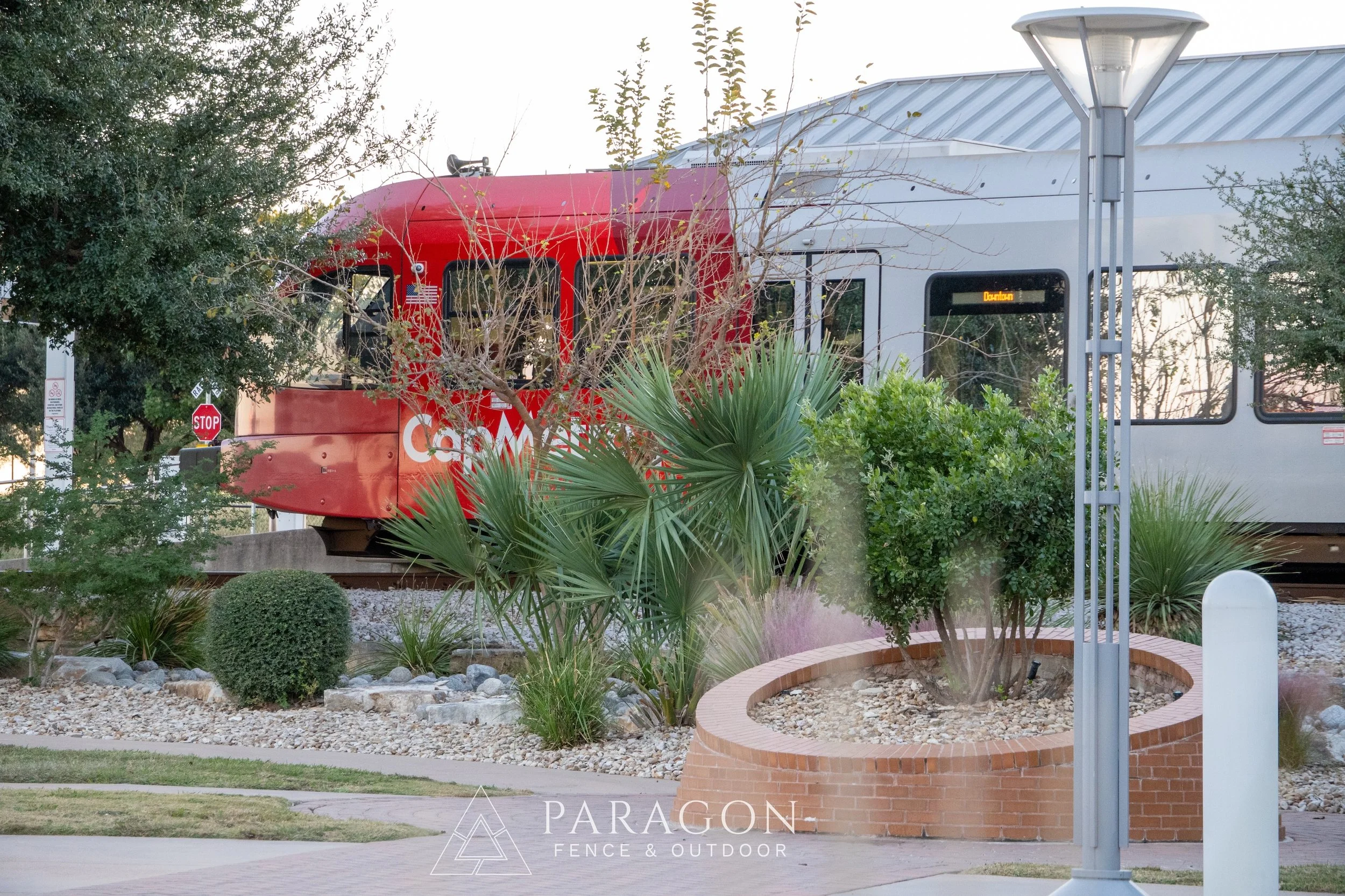 A train is passing by a landscaped area with trees, bushes, and a brick circular plant bed. The train is red and white, with a digital sign displaying 'Davenport,' and is partially obscured by the foliage. A streetlamp and a stop sign are visible nearby. The image is taken during daytime.