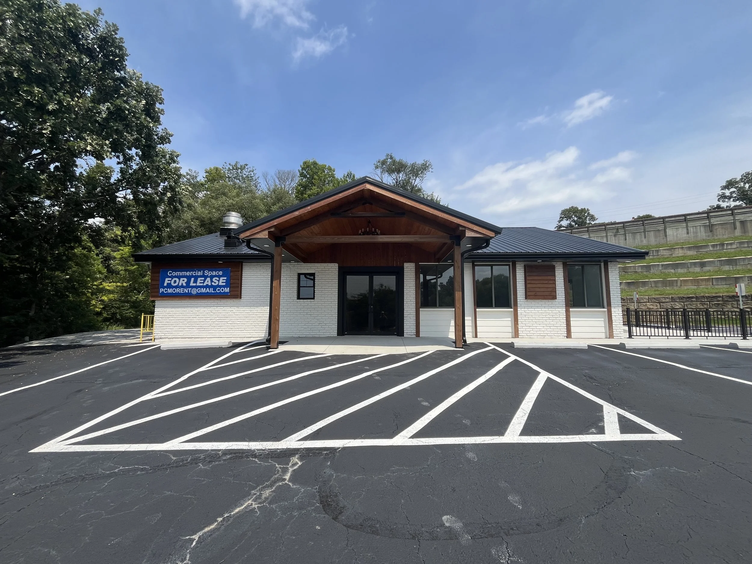 A small commercial building with a sign that reads 'For Lease', surrounded by parking spaces with freshly painted white lines, and a wooded area in the background.