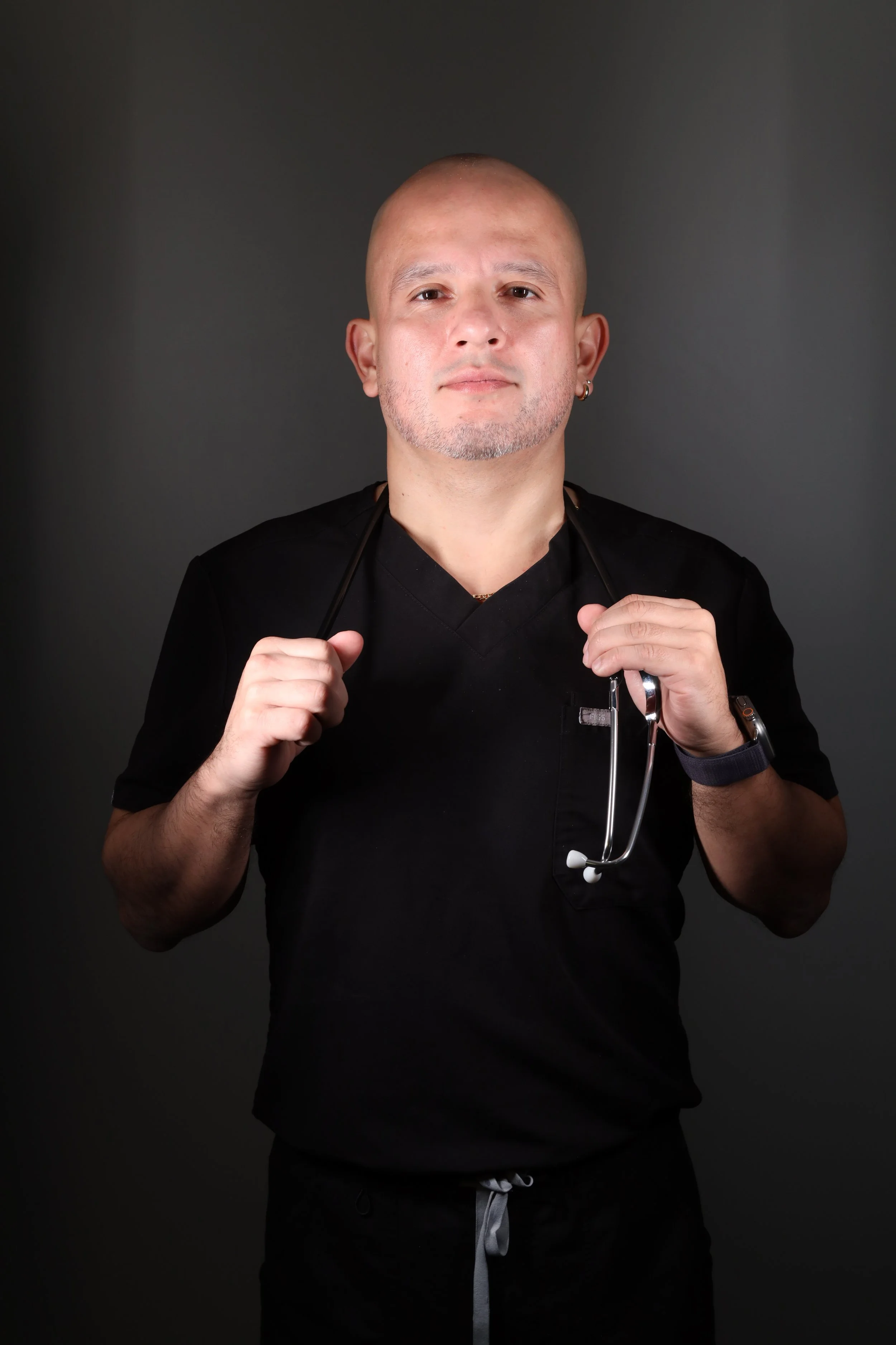 A male healthcare professional with a stethoscope around his neck standing against a dark background.