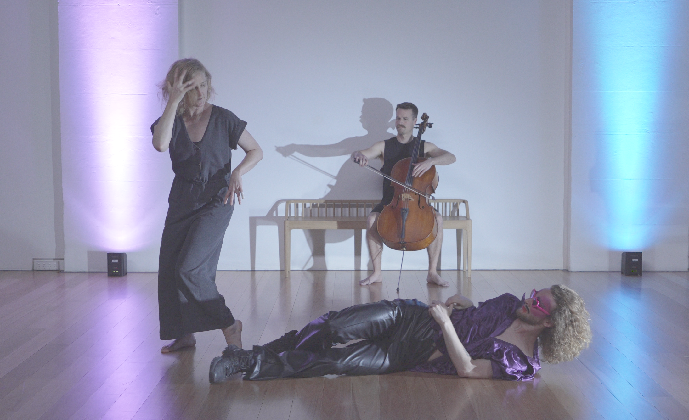 Three performers in a contemporary dance and music performance, with one woman standing in front, another woman lying on the floor, and a man sitting on a wooden bench playing a cello, all on a stage with colored lighting.