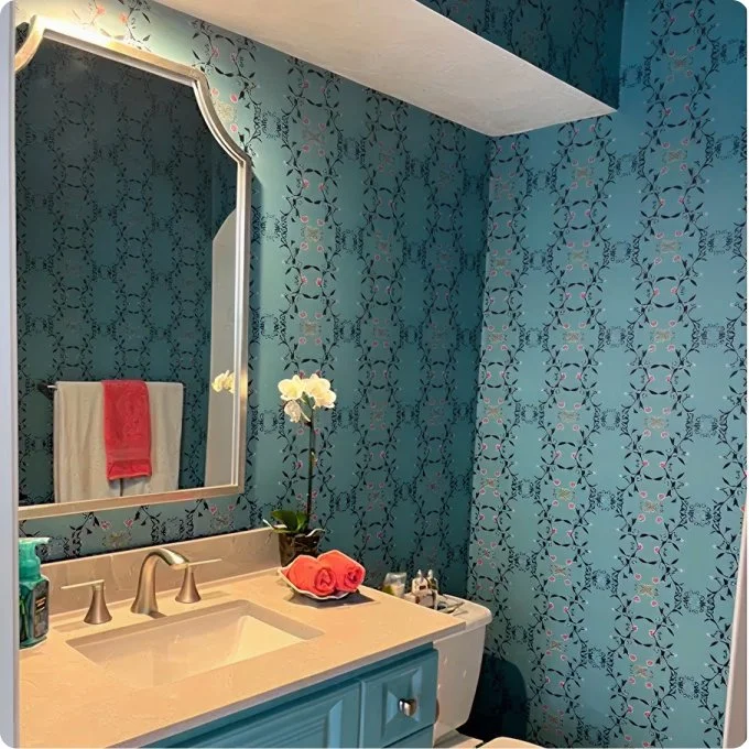 Bathroom with blue floral wallpaper, a mirror, a beige sink vanity with toiletries, and a towel with a red towel on a wall hook.
